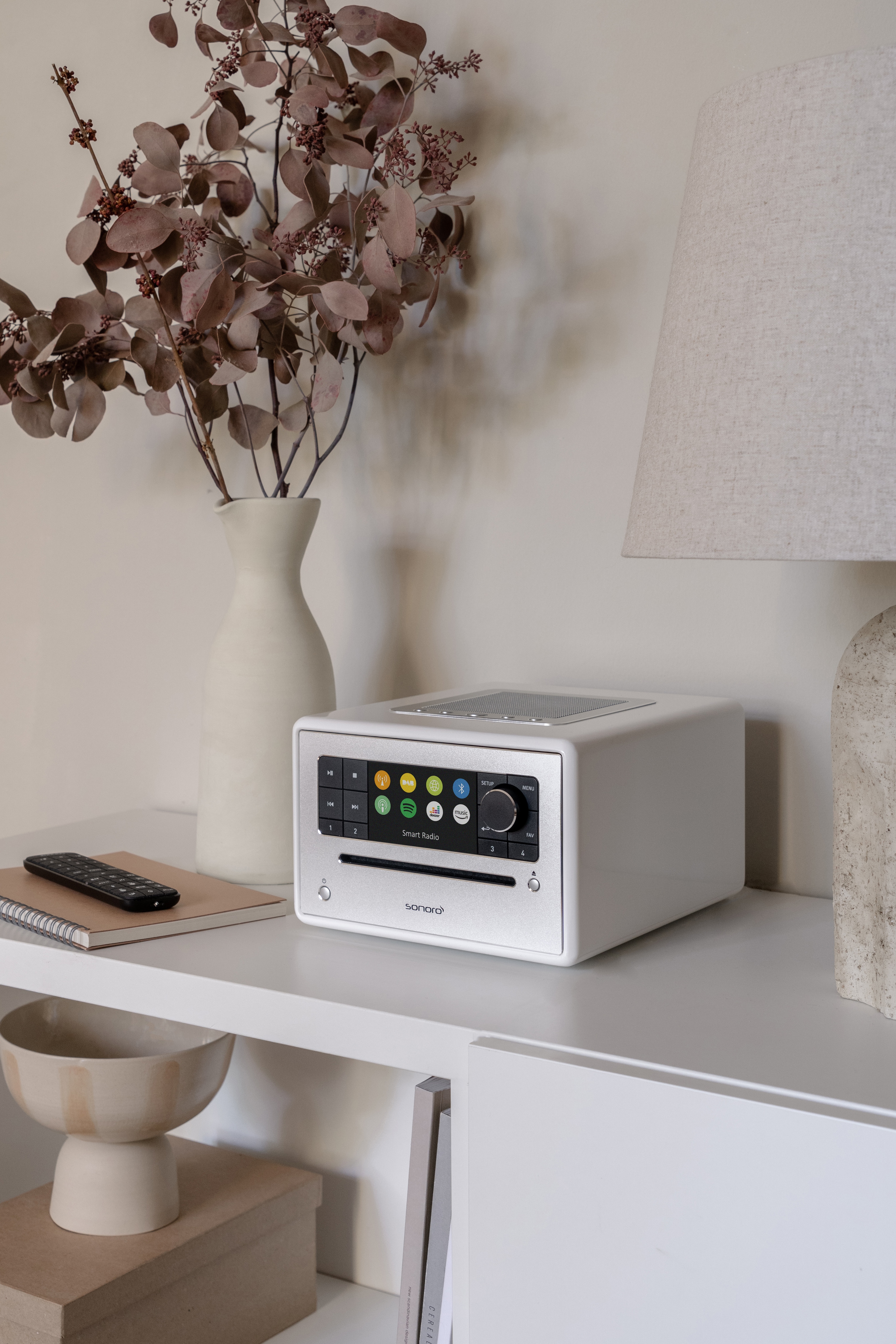 Witte stereo met een scherm op een plank. Vaas met bloemen en een lamp ernaast.