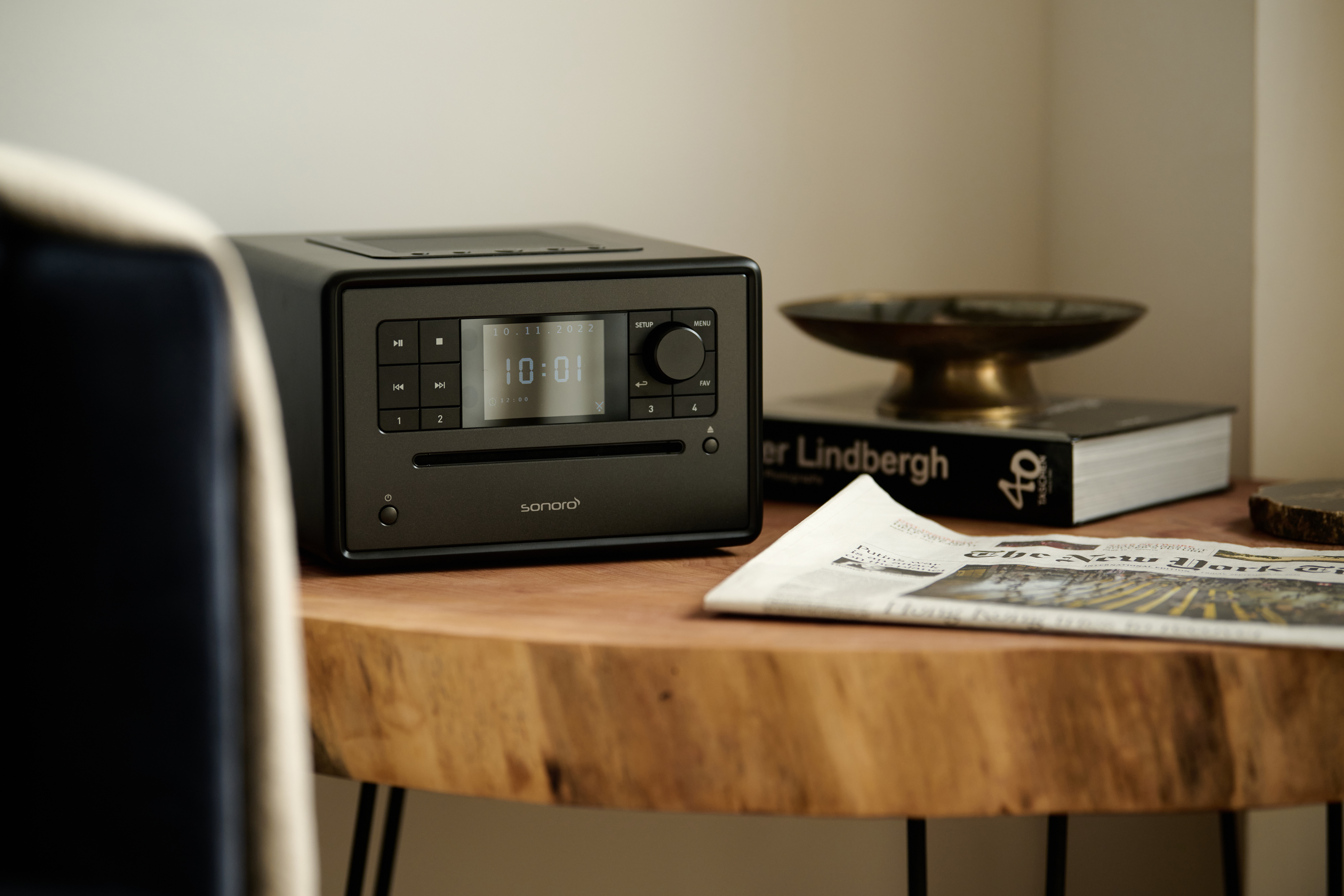 Donkergrijze slimme radio op een houten tafel met krant en een boek. Een stoel links.