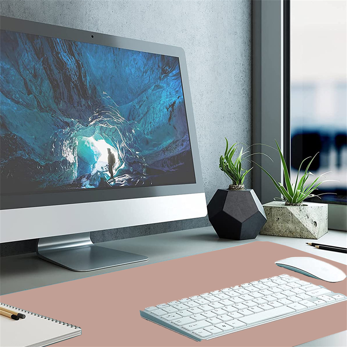 Arbeitsplatz-Setup: Computermonitor, Tastatur, Maus und Topfpflanzen auf einer pinkfarbenen Schreibtischunterlage.