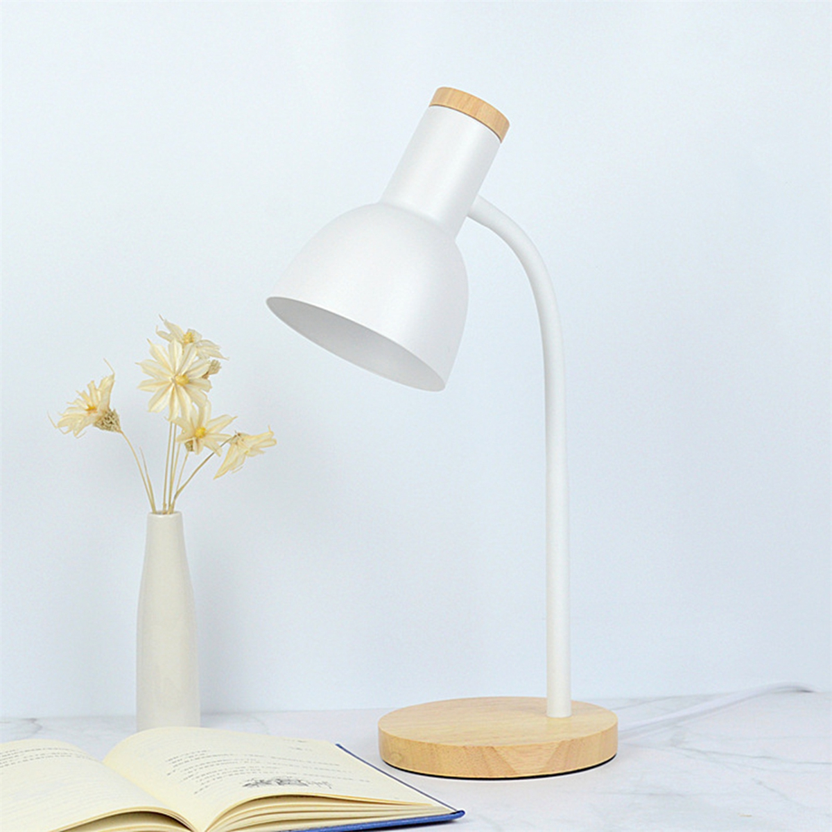 Eine weiße Schreibtischlampe mit Holzsockel und eine Vase mit Trockenblumen neben einem offenen Buch.