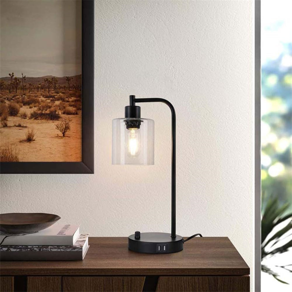 Schwarze Schreibtischlampe mit transparentem Glasschirm. Auf einem dunklen Holztisch. Weiße Wand im Hintergrund.