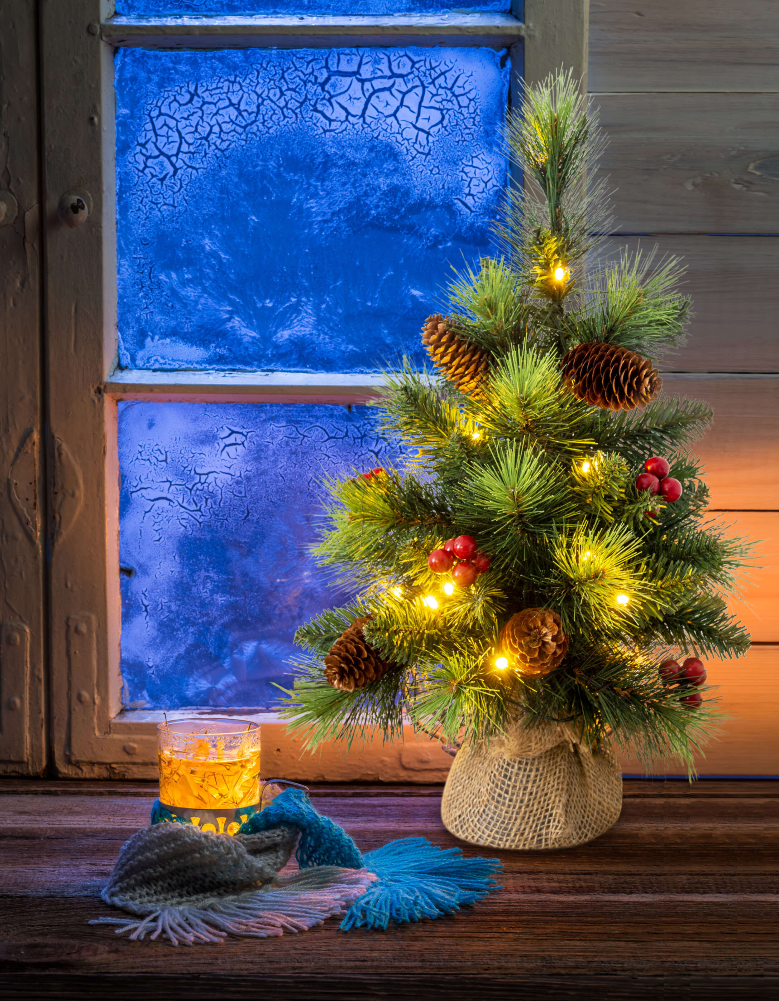 Ein kleiner Weihnachtsbaum mit Lichtern, Tannenzapfen und Beeren neben einem Fenster mit gefrorener Aussicht.
