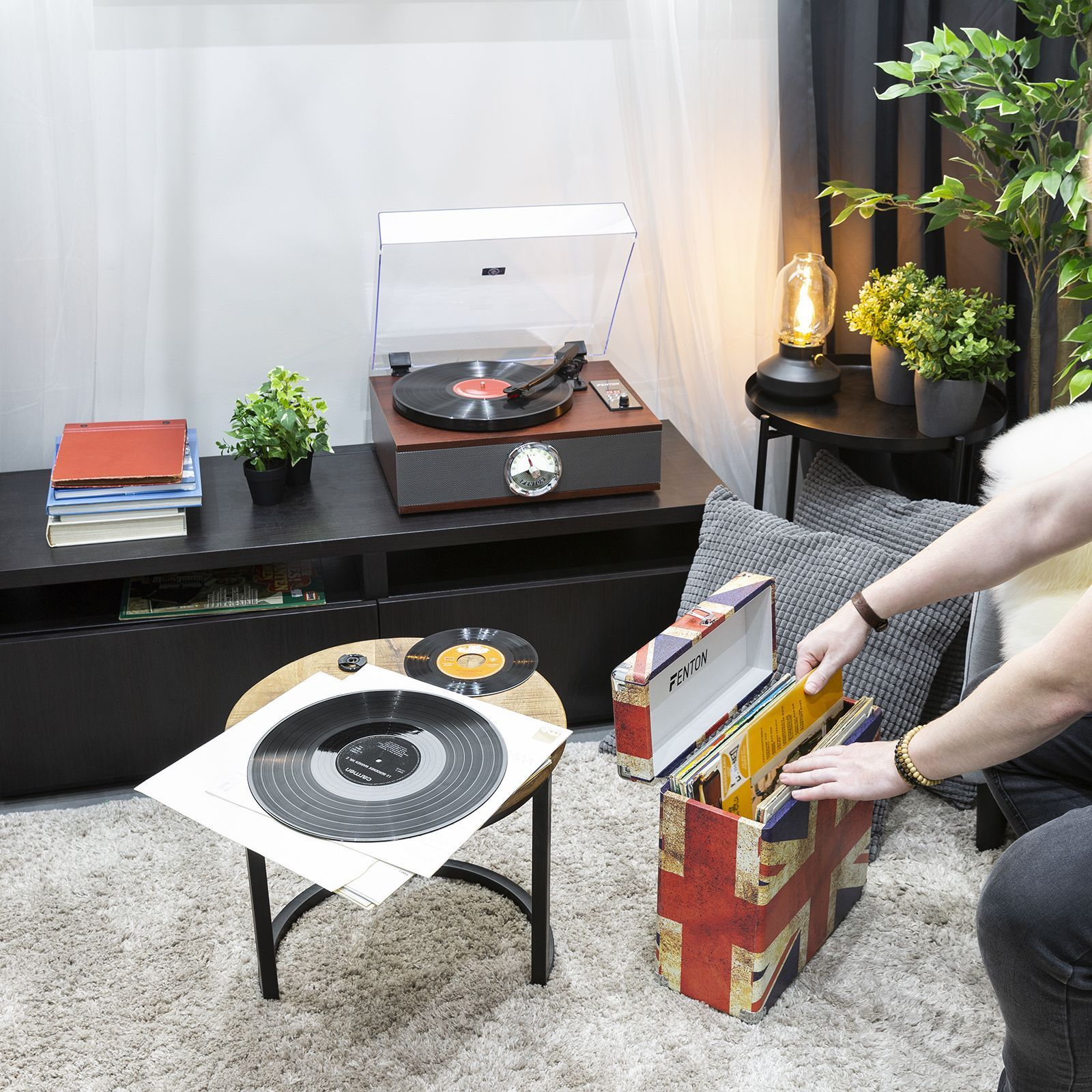 Eine Person baut einen Plattenspieler mit Schallplatten auf. Es gibt Pflanzen und eine Lampe.