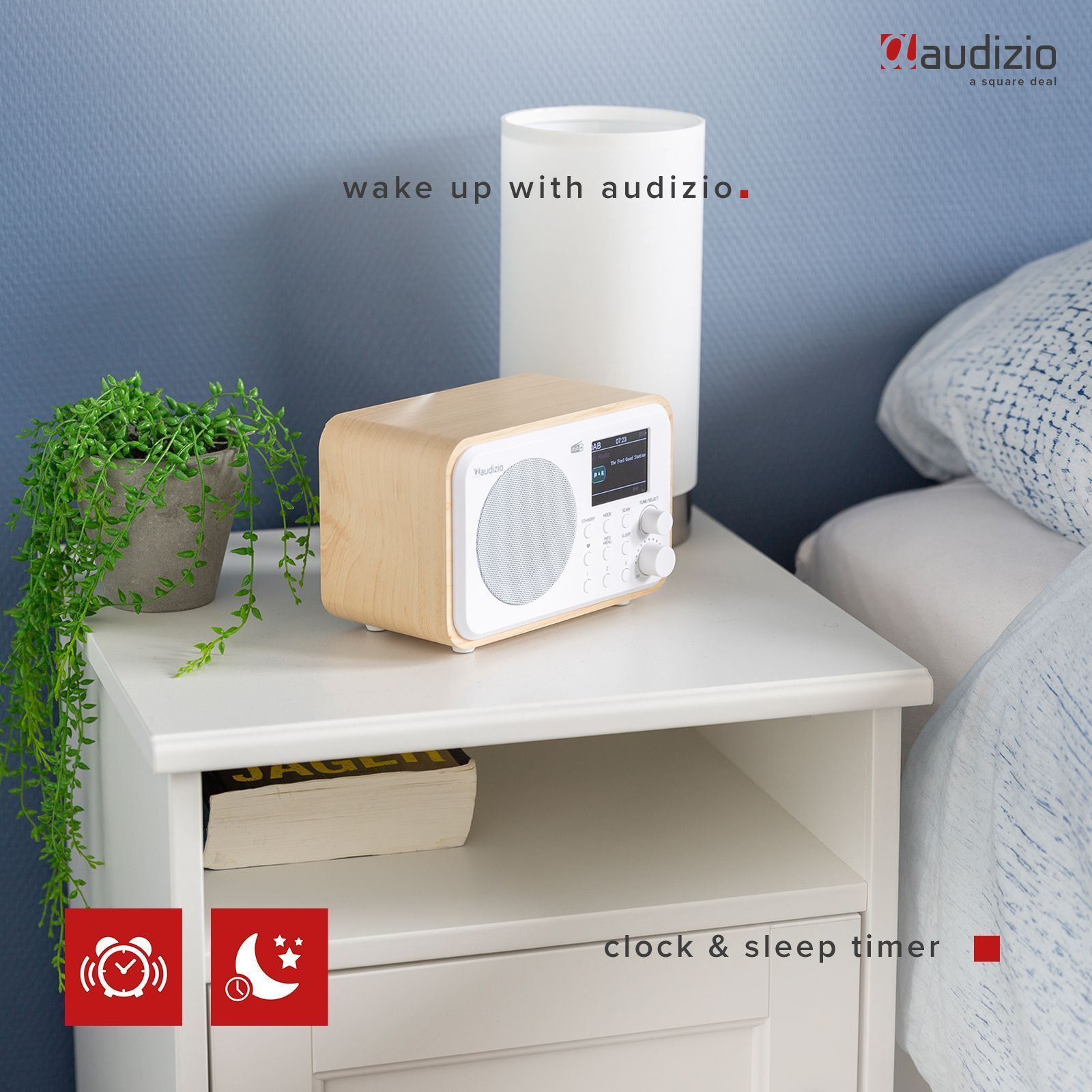 Radio de madera en mesita de noche blanca con lámpara, planta y libro, fondo de pared azul.