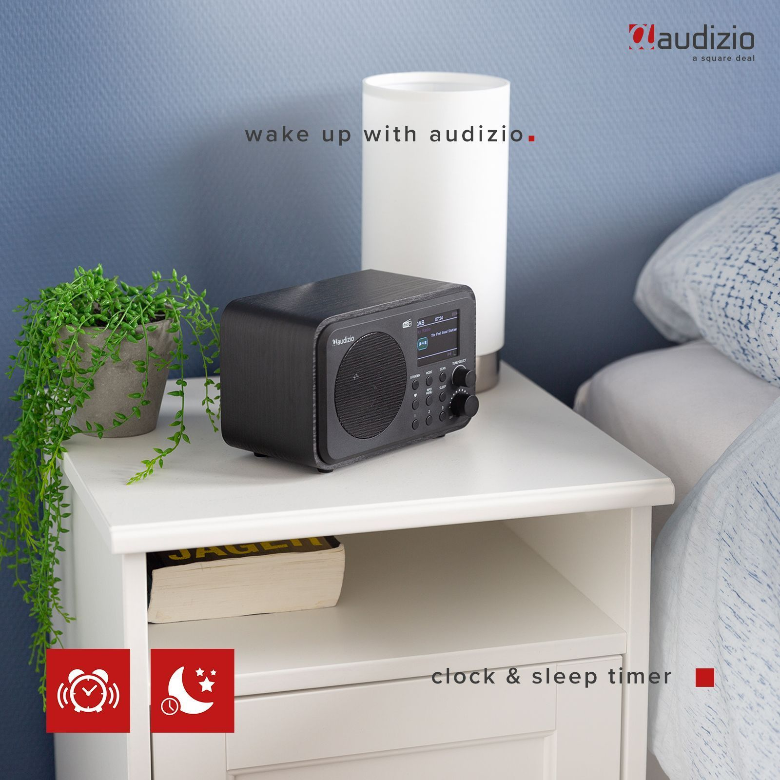 Una radio negra sobre una mesita de noche blanca con lámpara, planta y libro. Fondo de pared azul.