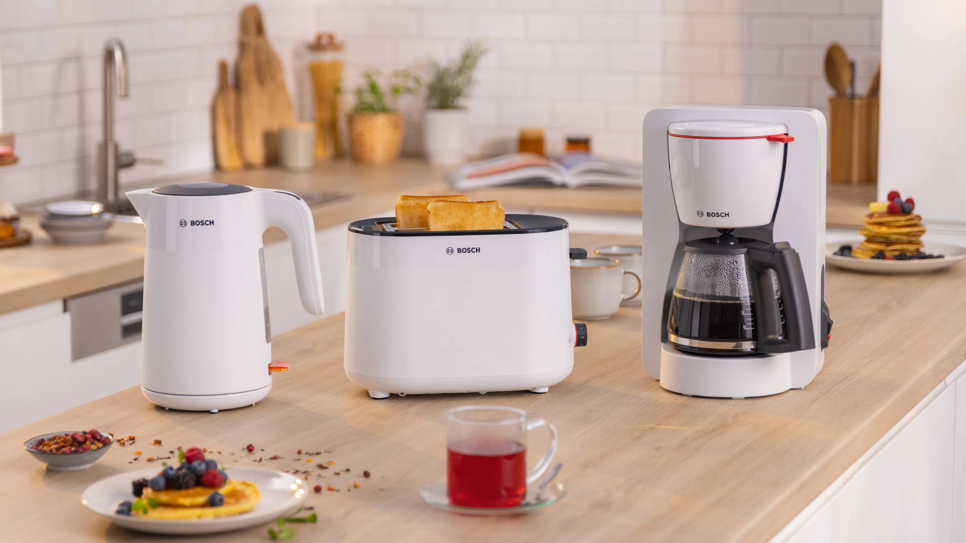 Biały czajnik, toster i ekspres do kawy BOSCH na drewnianym blacie z jedzeniem.