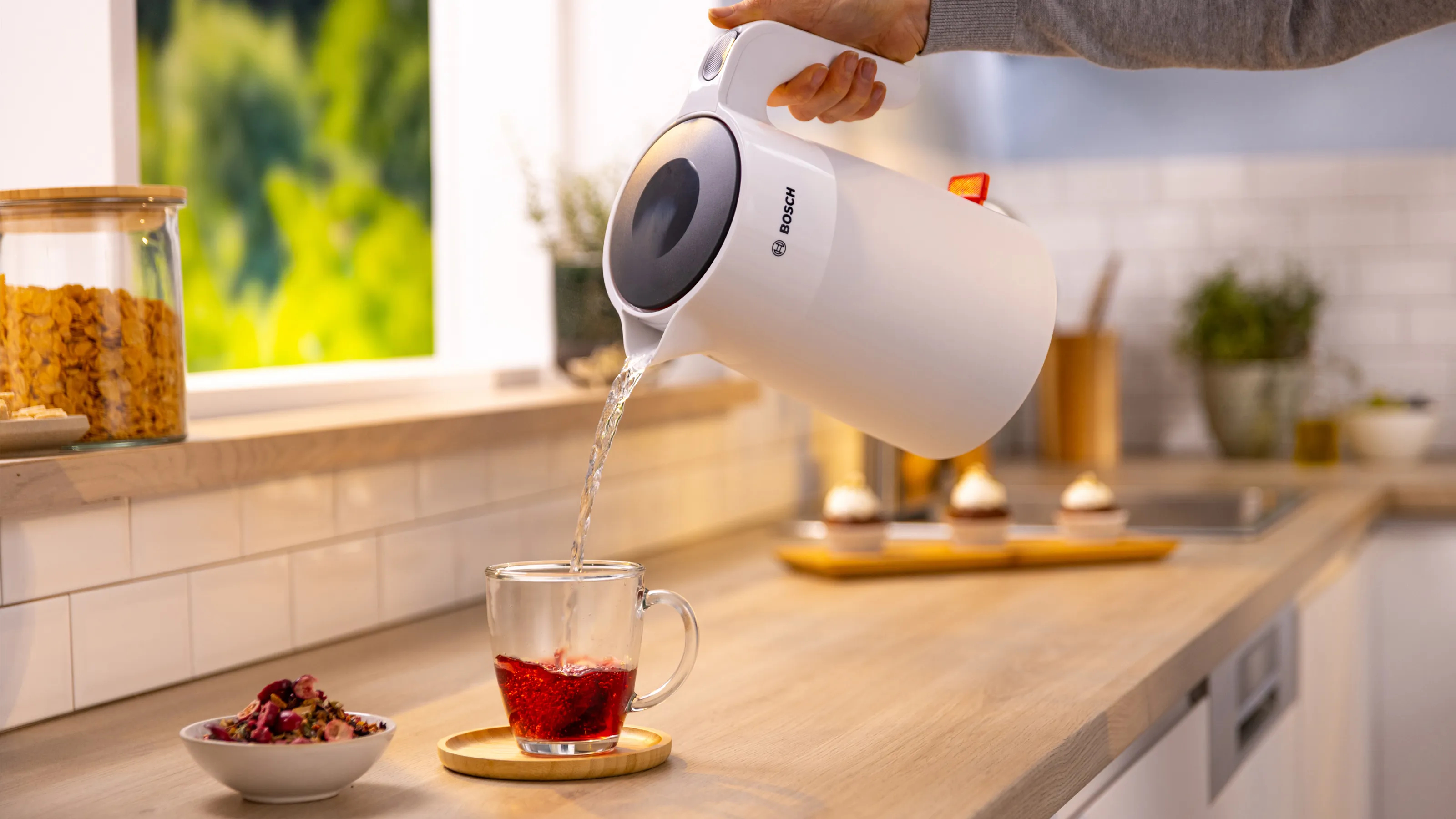 Dłoń wlewa wodę z białego czajnika do szklanki. Kuchnia, drewniany blat i babeczki.