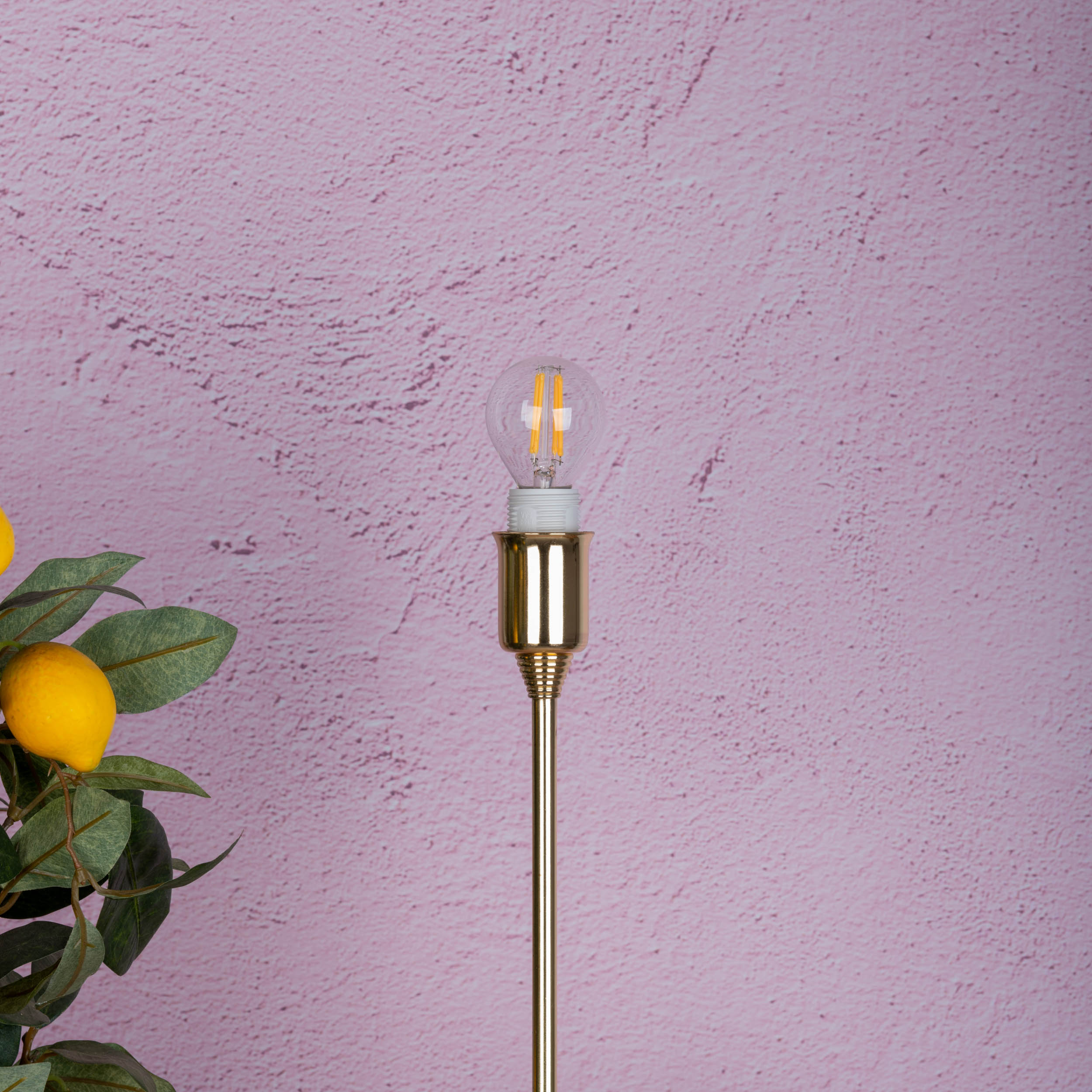 Eine Lampe mit einer Glühbirne auf einem rosa strukturierten Hintergrund. Zitronen und Blätter sind links unten.
