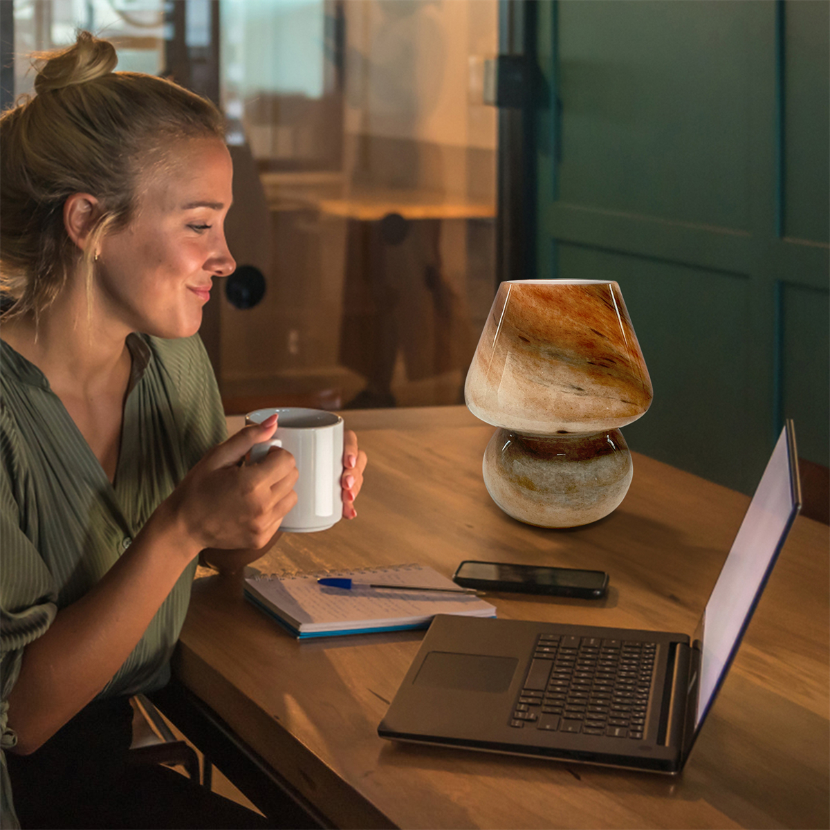 Eine Frau trinkt aus einem weißen Becher, mit einer Lampe und einem Laptop auf einem Holztisch.