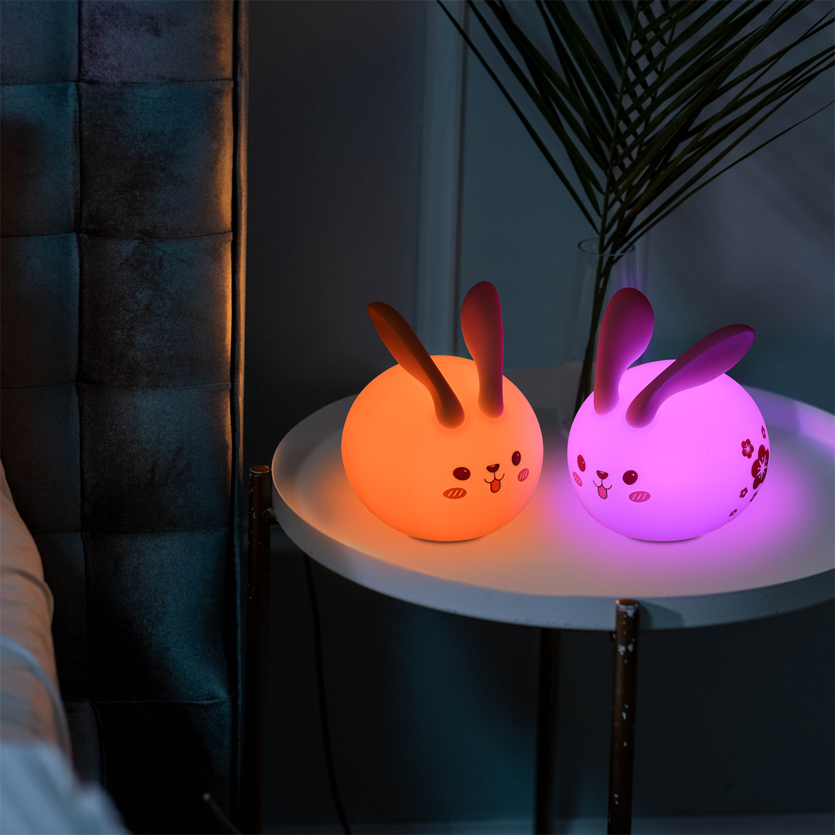 Zwei hasenförmige Lampen, eine orange und eine rosa, mit einem weißen Tisch und dunklem Hintergrund.