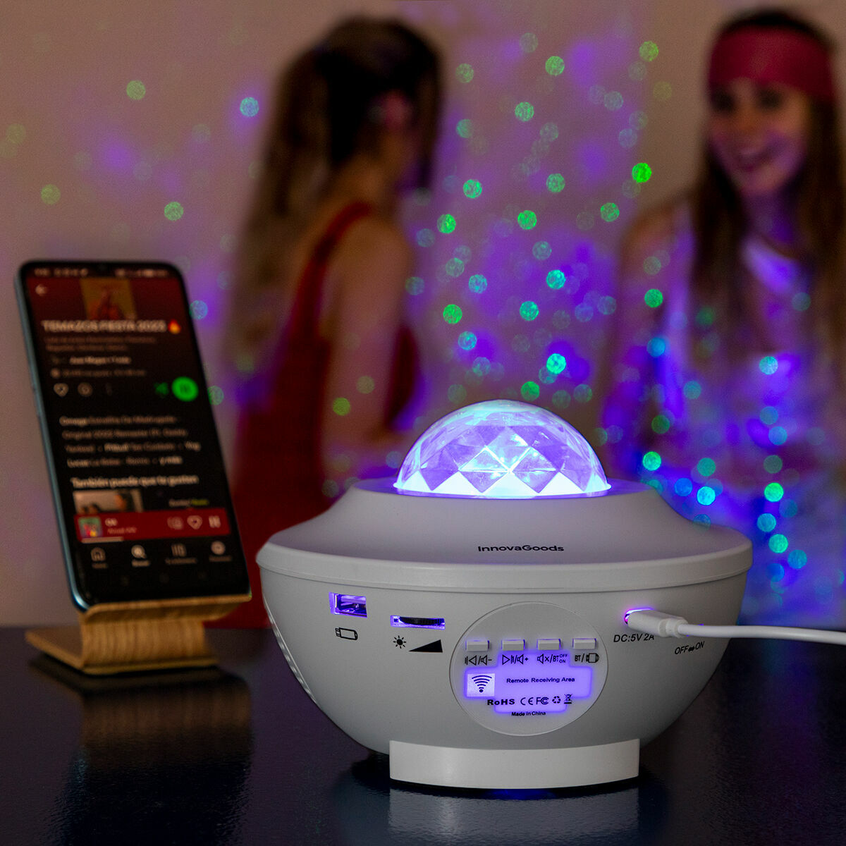 Ein weißer Galaxieprojektor mit einem Telefon und zwei verschwommenen Figuren im Hintergrund.