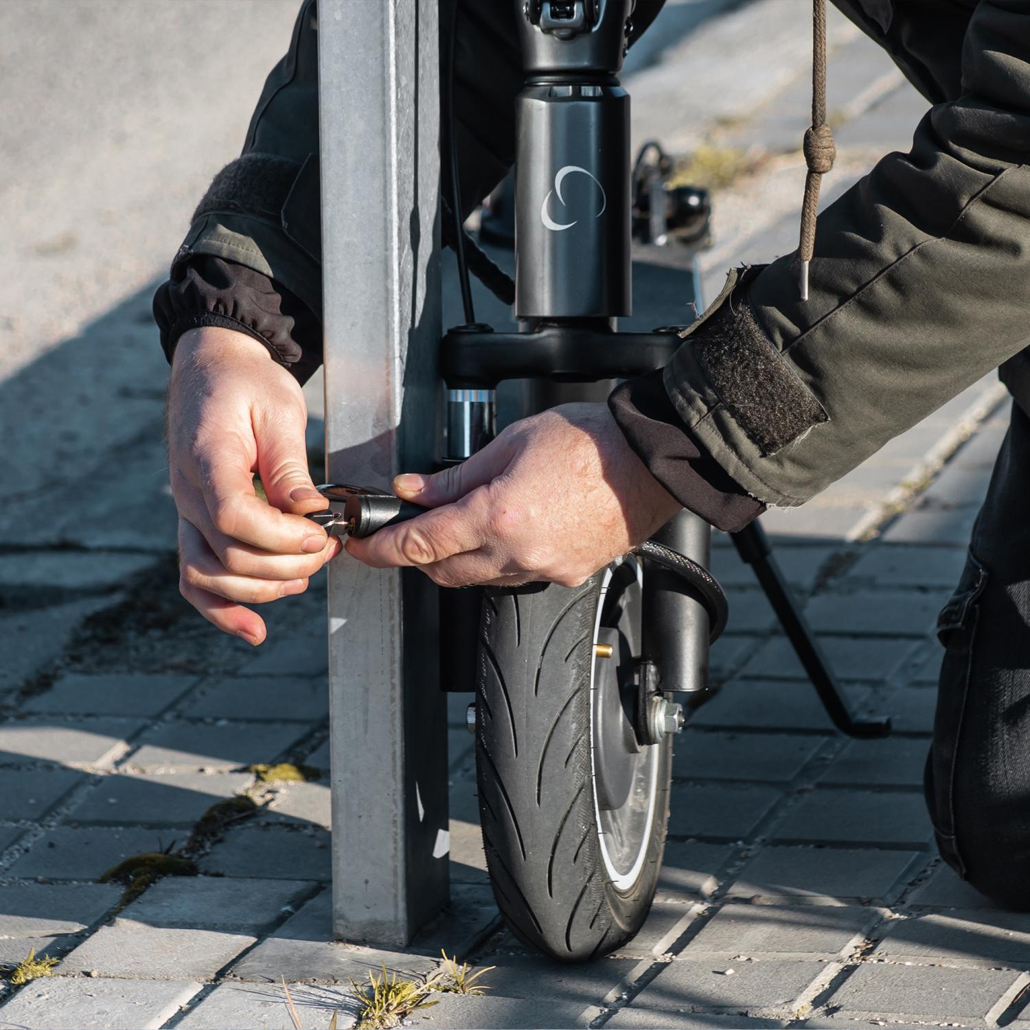 Una persona usando una llave para asegurar un scooter eléctrico a un soporte metálico en una superficie pavimentada.
