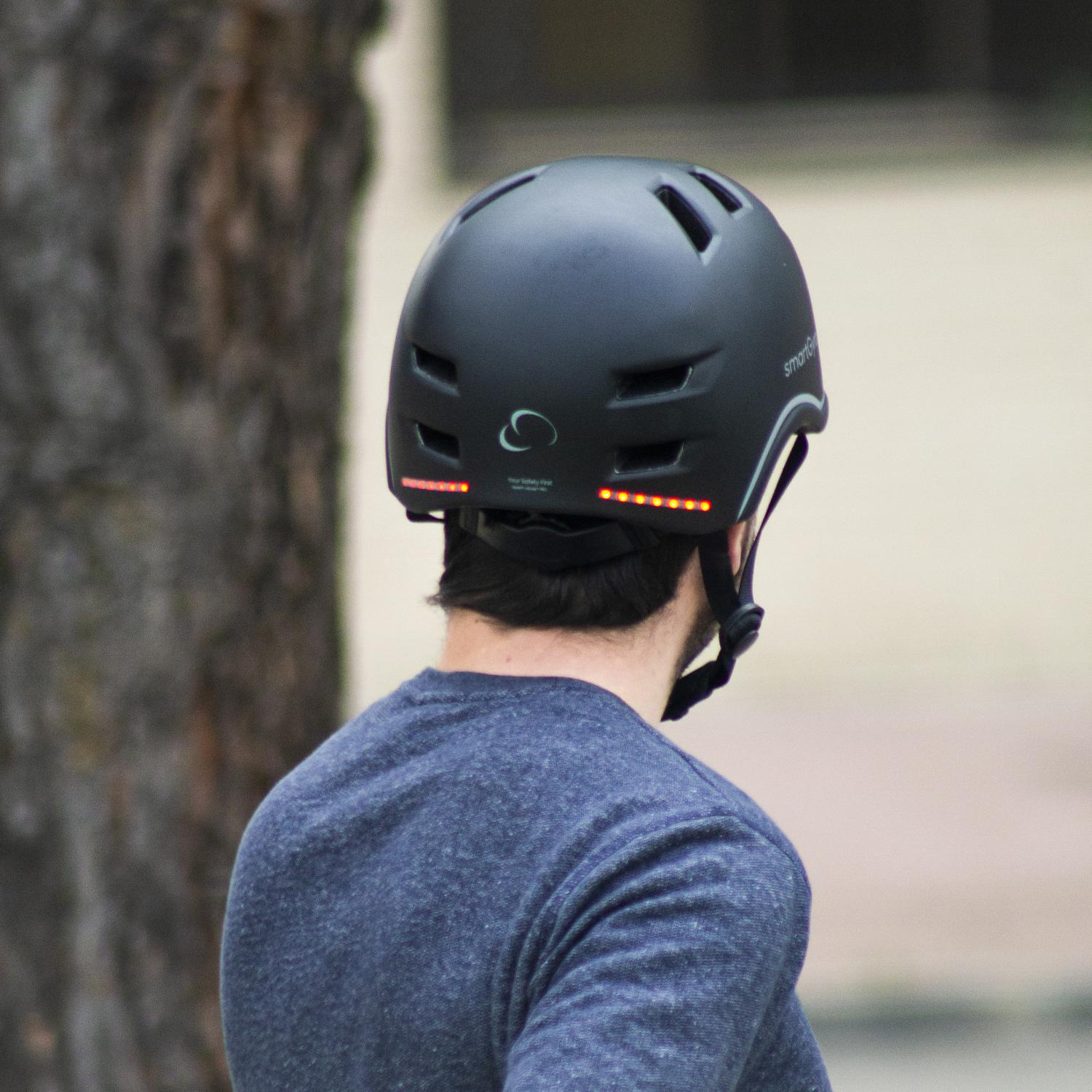 Una persona con un casco de bicicleta negro con luces rojas en la parte trasera, de pie frente a un árbol.