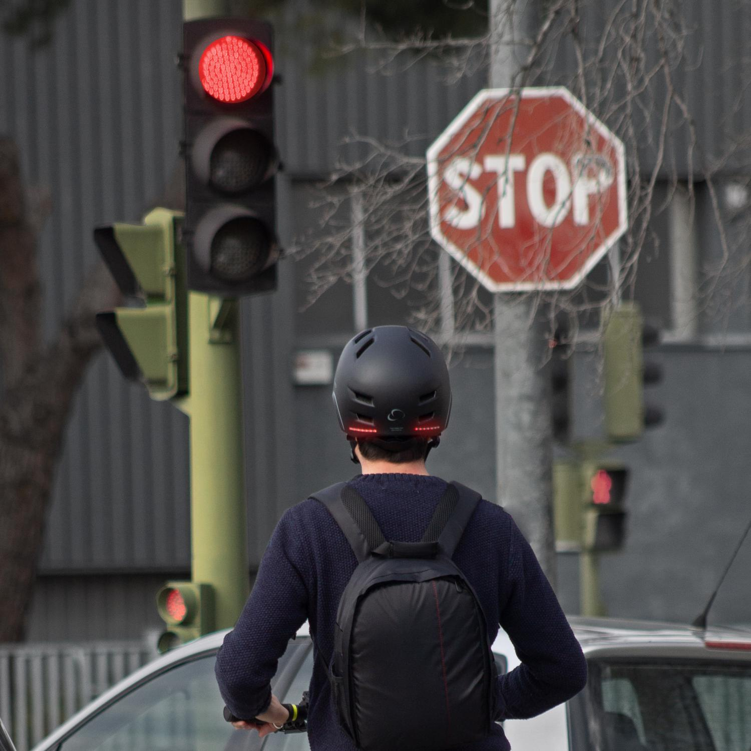 Persona en scooter hacia semáforo rojo y señal de stop. Con casco y mochila.