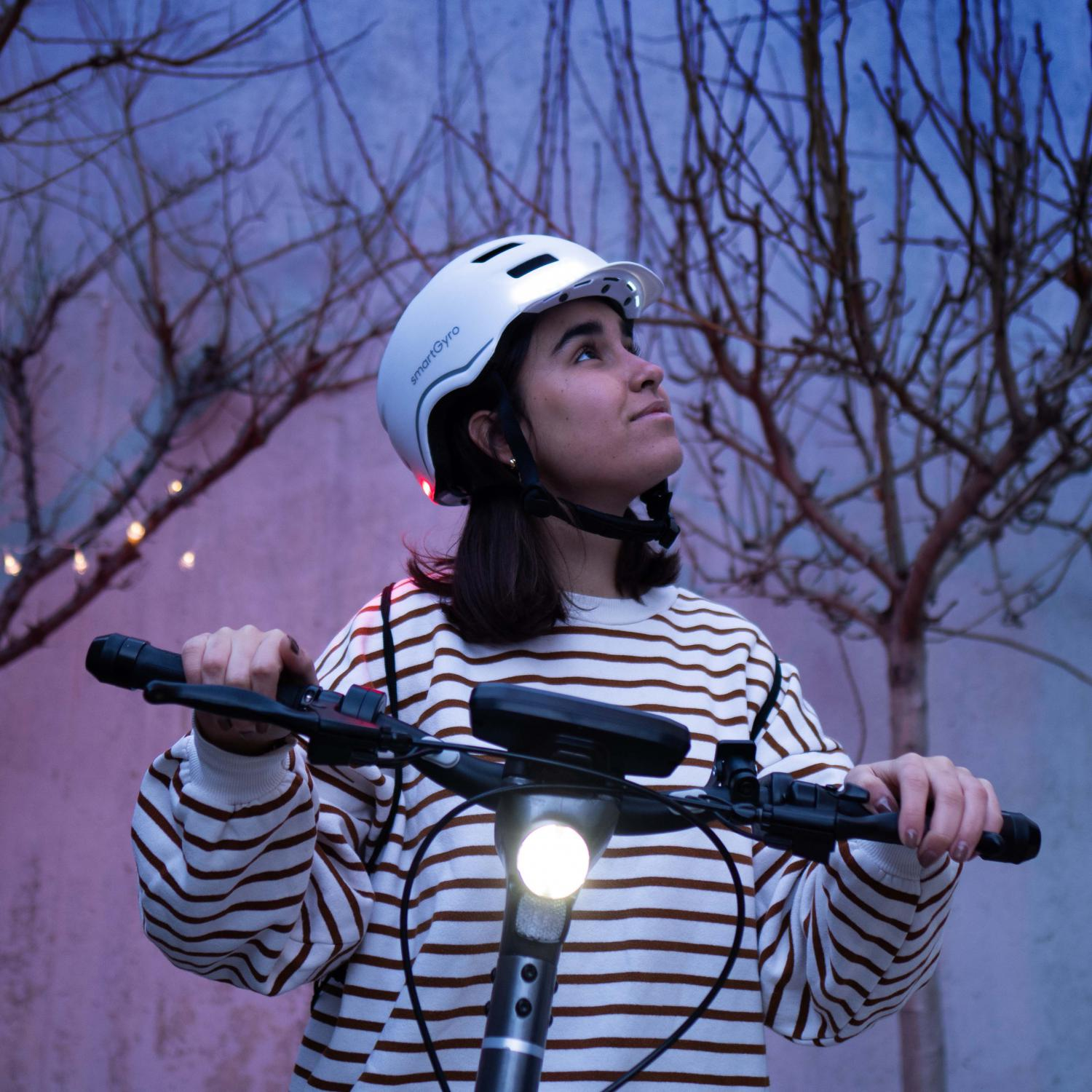 Mujer con casco blanco con luces, sosteniendo el manillar de un scooter. Fondo: árboles.