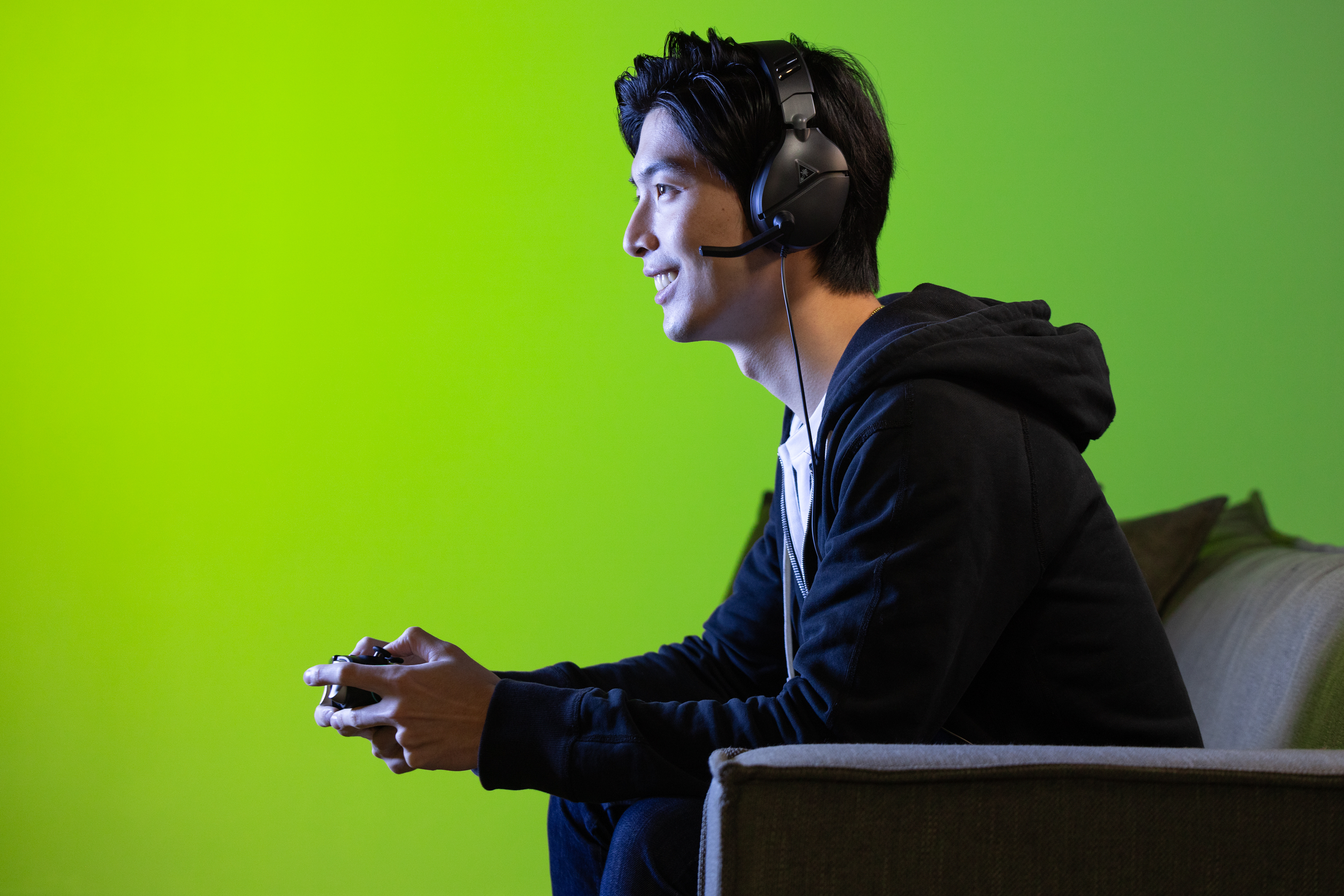 Hombre jugando en un sofá con auriculares y mando, fondo verde.