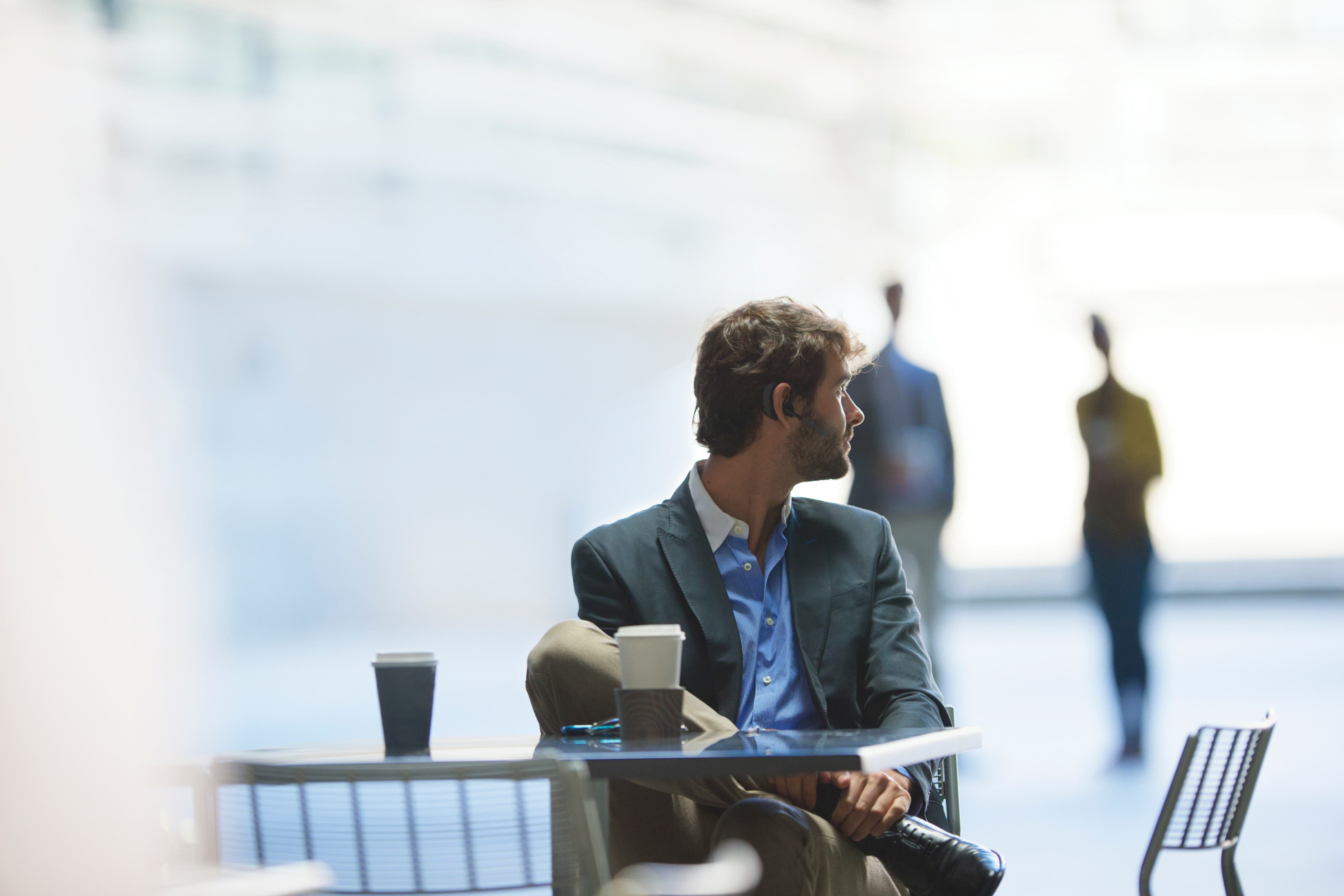 Mężczyzna w garniturze siedzi przy stole patrząc w prawo. Na stole dwie filiżanki kawy. Sylwetki w tle.