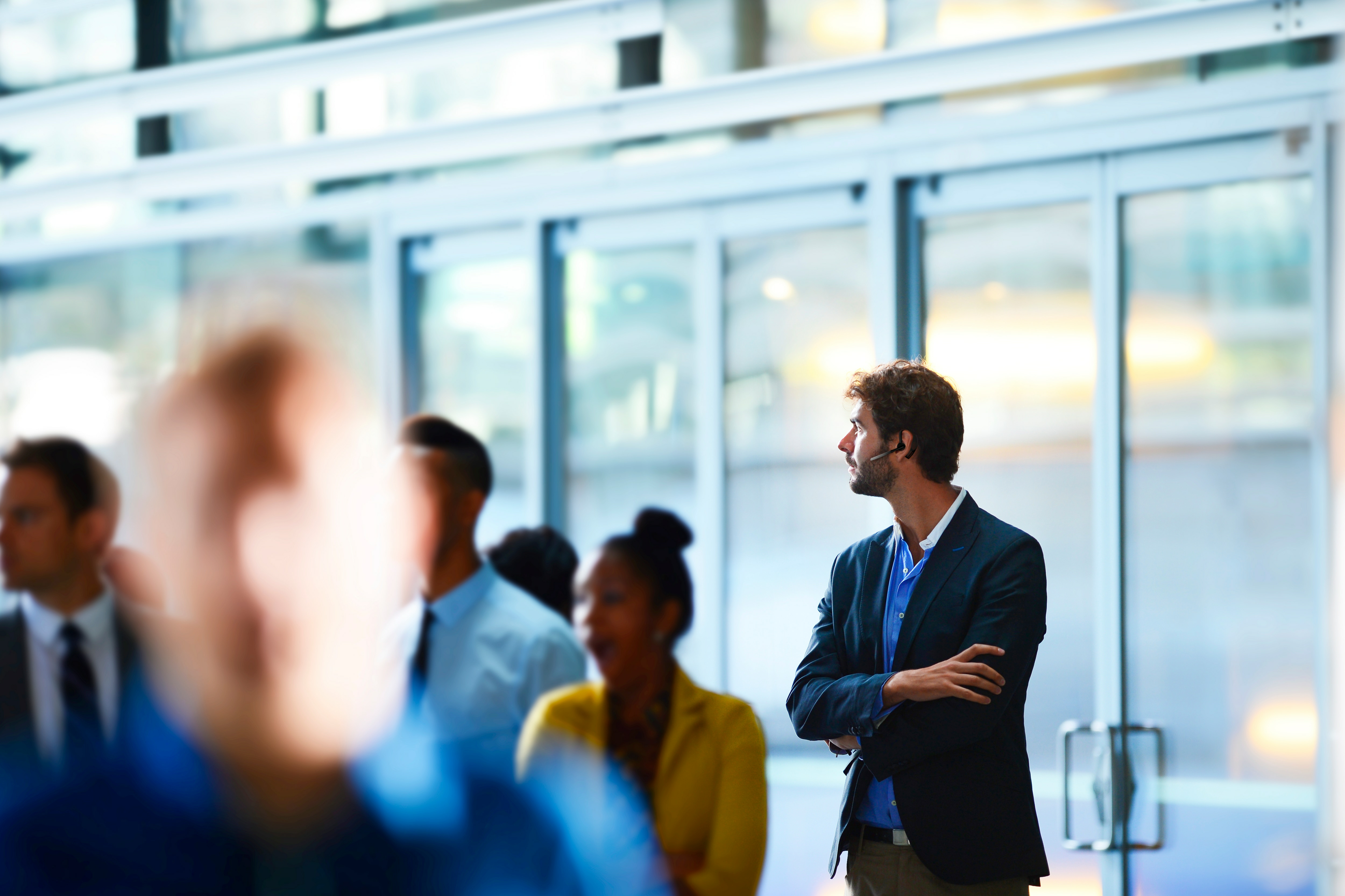 Mężczyzna w słuchawce, stojący w tłumie. Tłem jest szklany budynek z wieloma ludźmi.