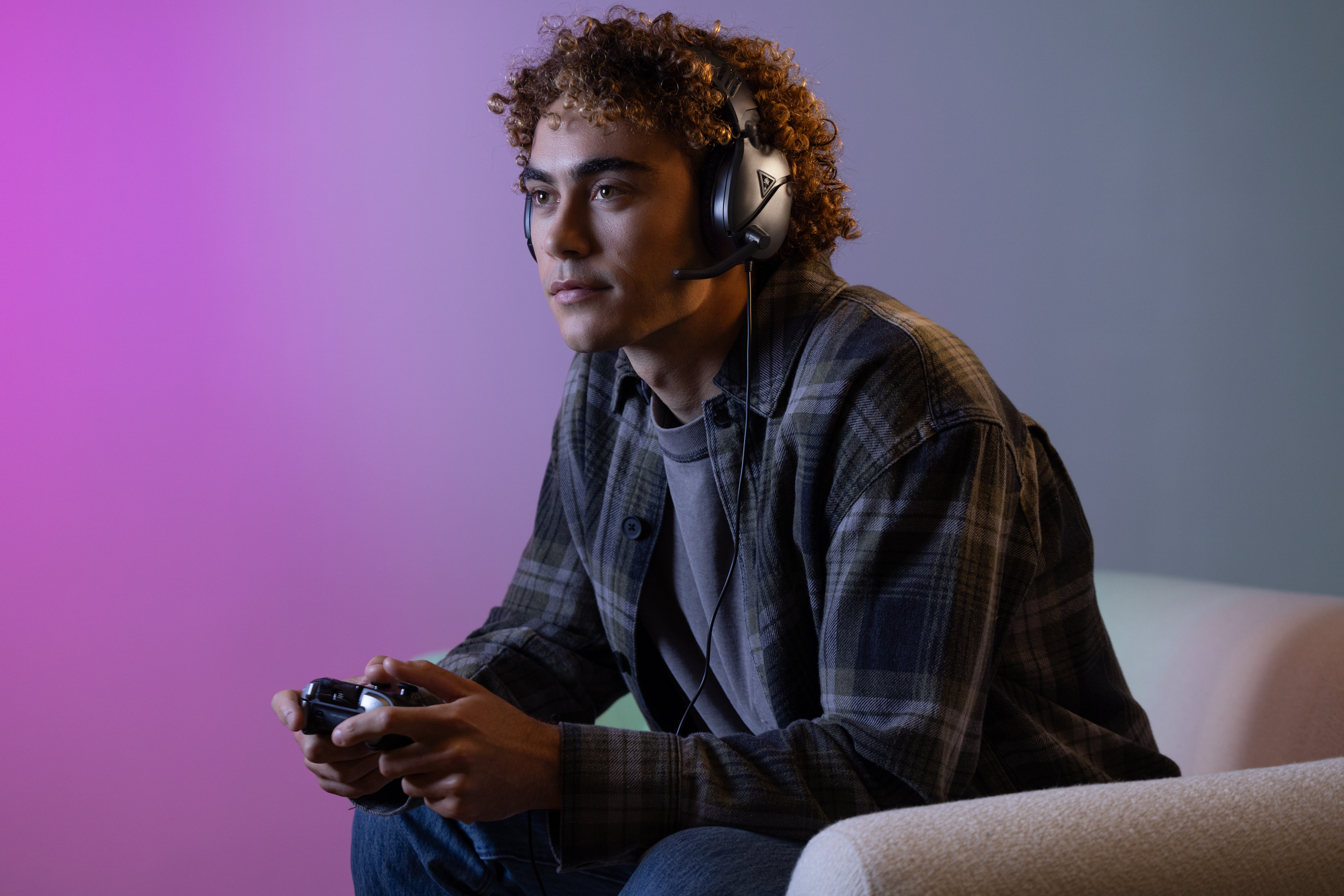 Młody mężczyzna w słuchawkach do gier, trzymający kontroler i patrzący na ekran, siedzący na kanapie.