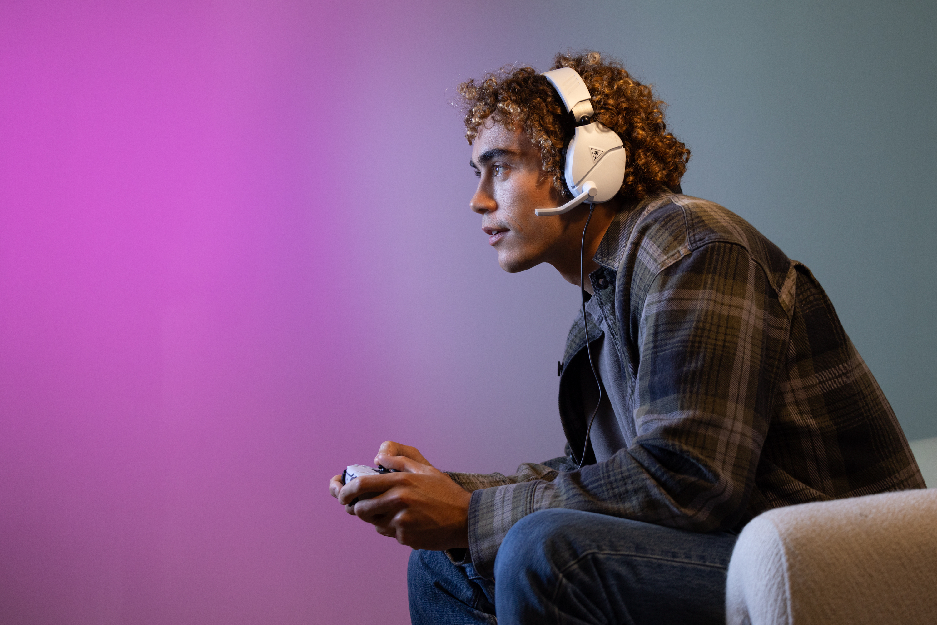 A young man with headphones playing a game, holding a controller, with a pink and teal background.