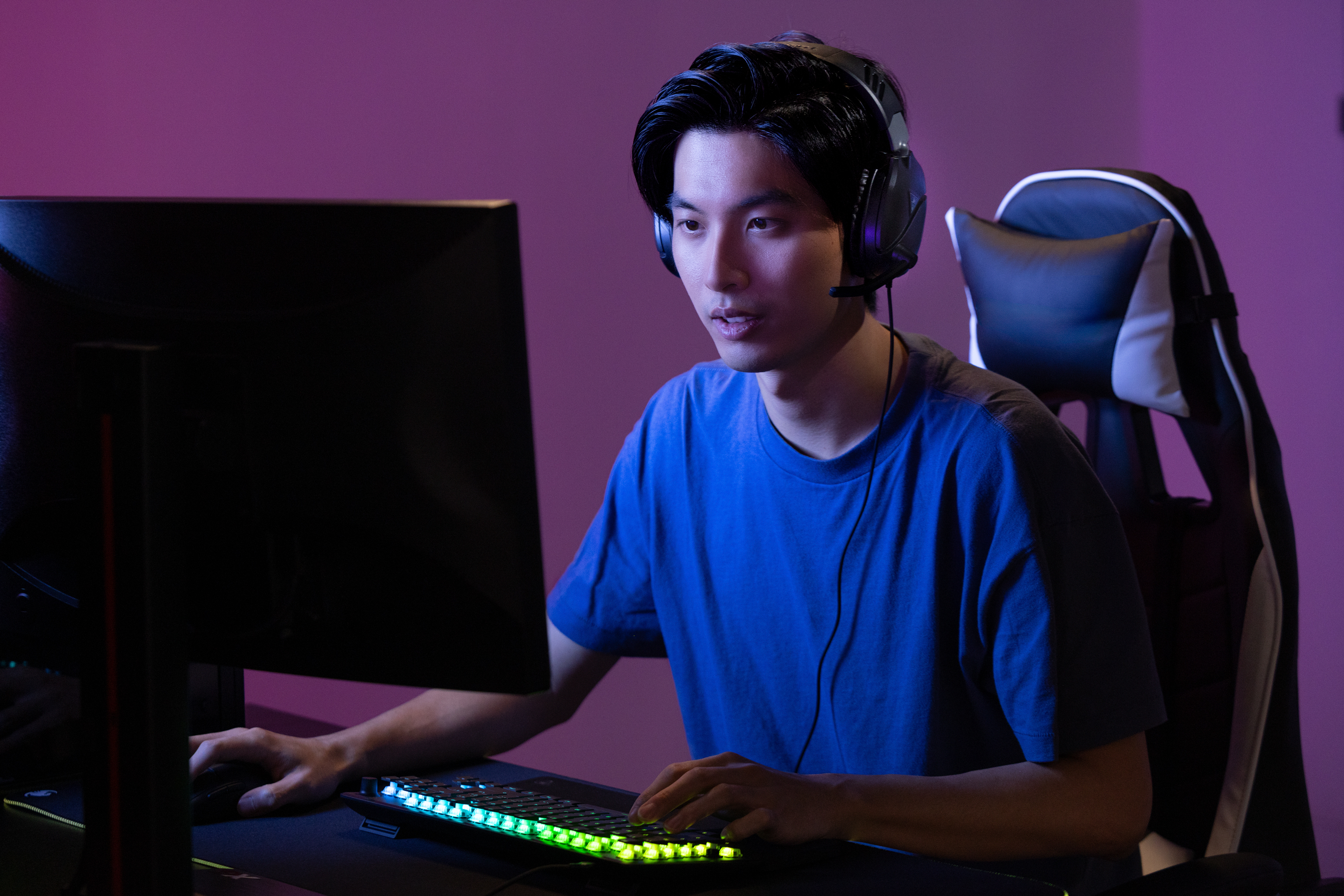 Mężczyzna gra, ma na sobie zestaw słuchawkowy. Siedzi przy komputerze, klawiatura ze światłami, fioletowe i niebieskie światła.