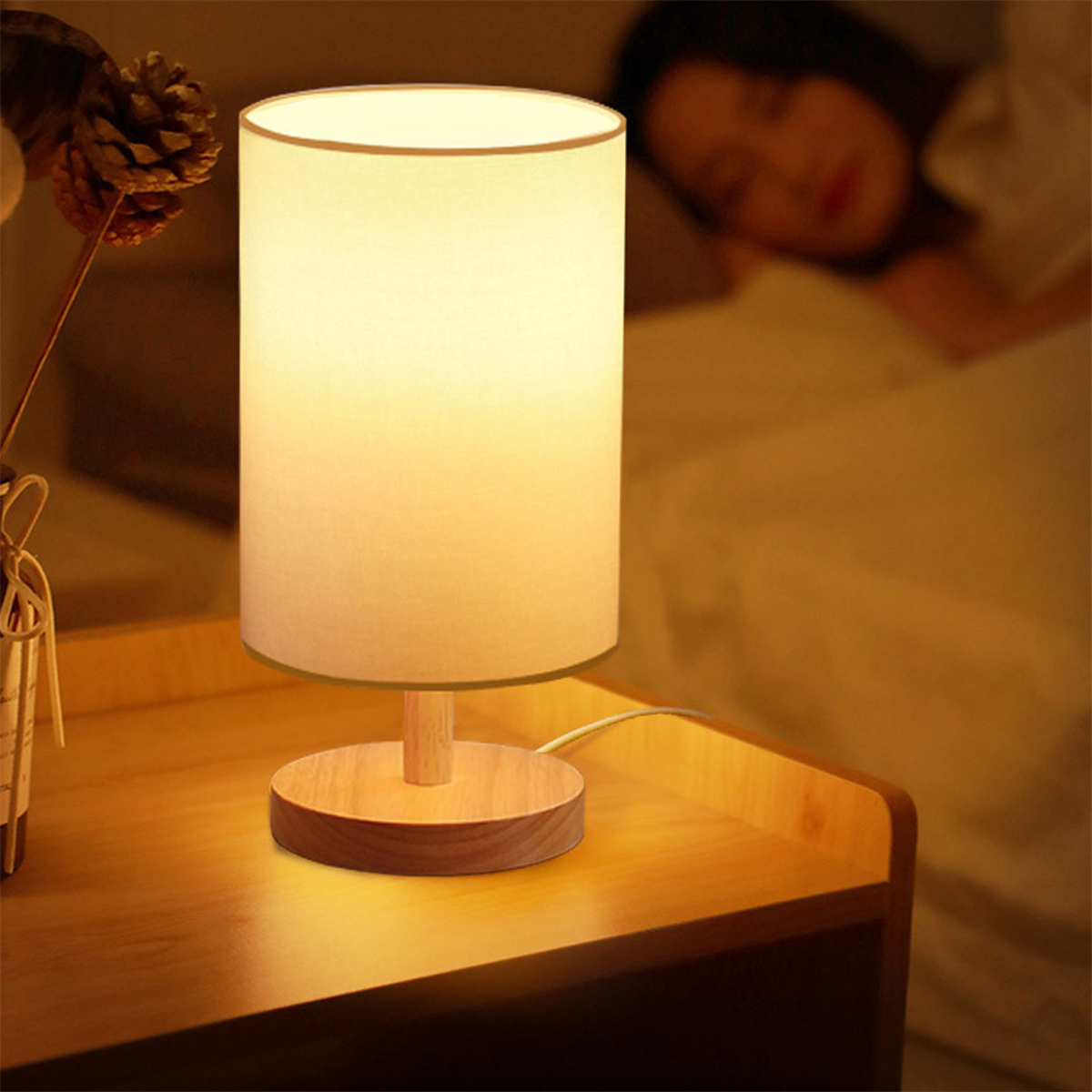 Tischlampe mit weißem Schirm auf Holzsockel, neben einer schlafenden Person im Bett.