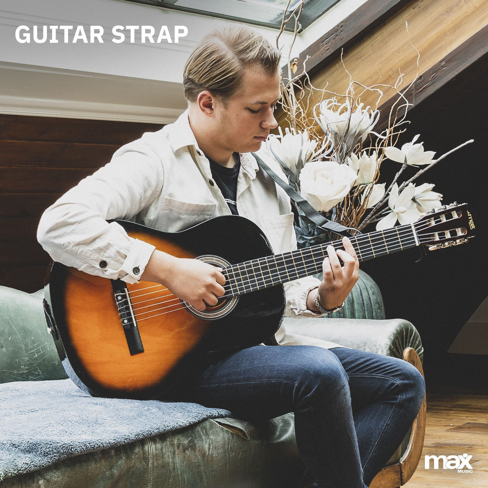 Ein Mann spielt Gitarre. Er trägt ein helles Hemd und Jeans, sitzt auf einem Sofa. Es gibt einen Gitarrengurt.