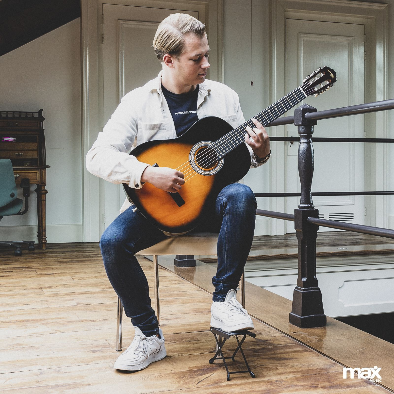 Ein Mann spielt Gitarre, während er auf einem Stuhl sitzt, mit Holzboden im Hintergrund.
