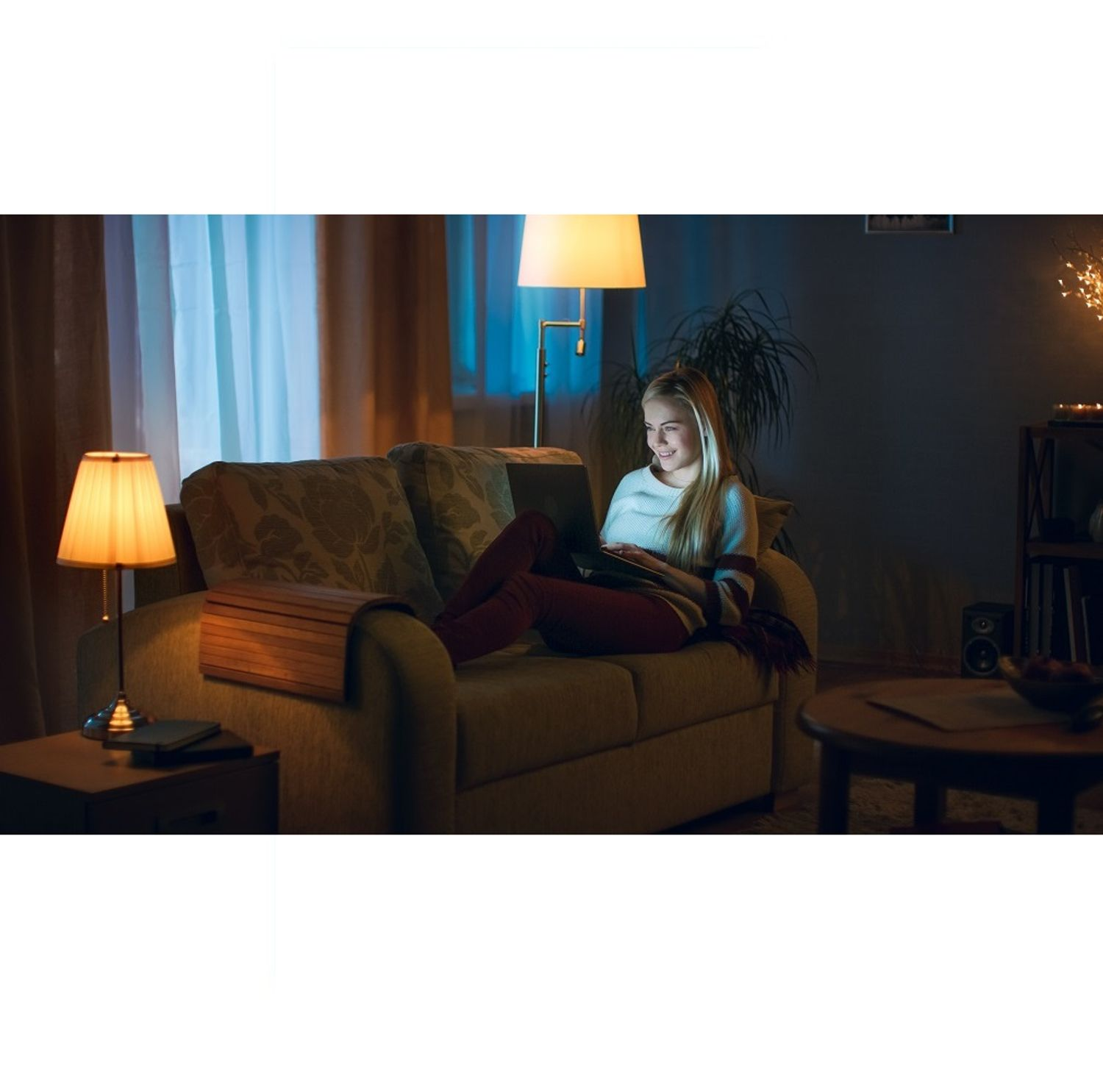 Kobieta na kanapie korzysta z laptopa w nocy. Lampy i rośliny w tle.