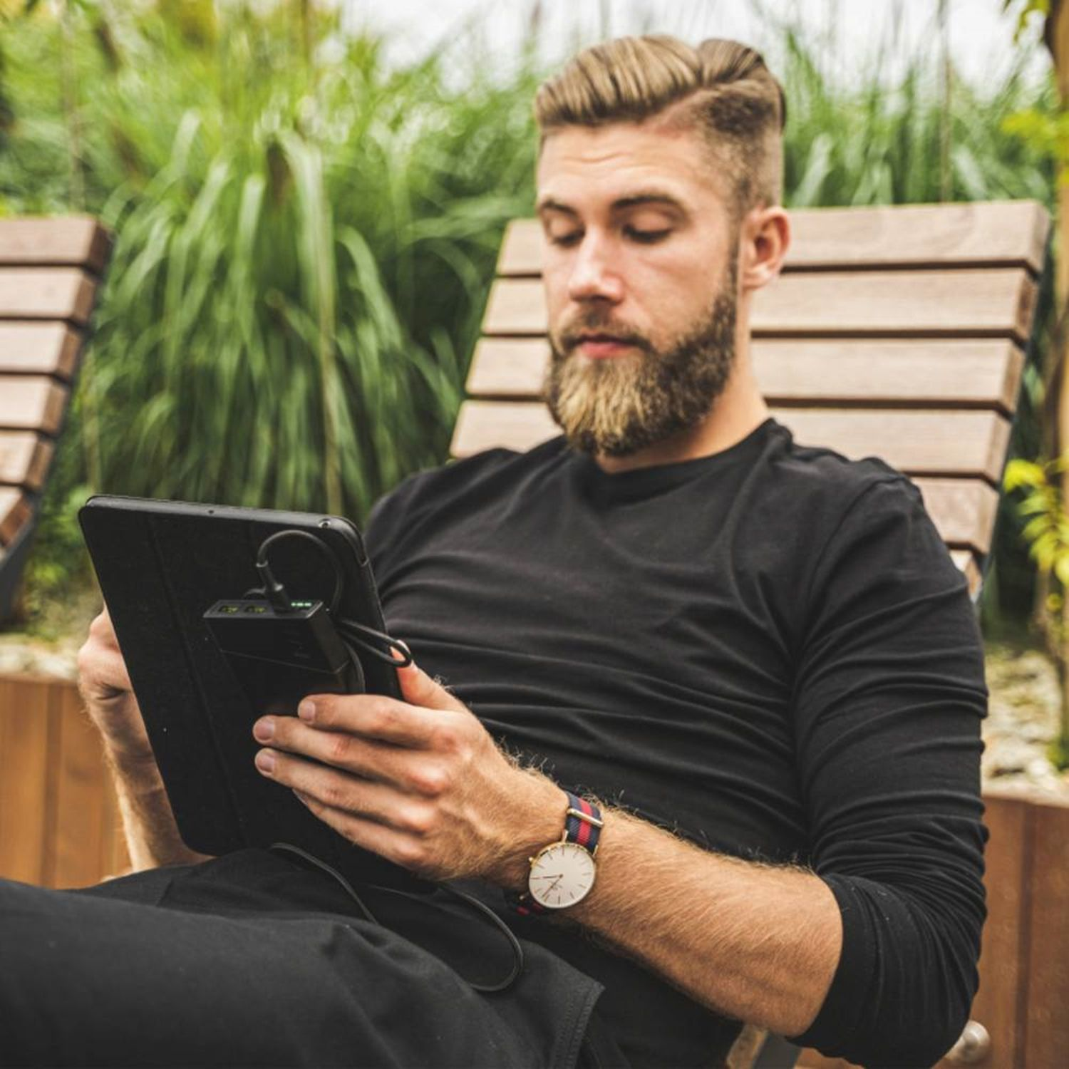 Ein Mann, der im Freien ein Tablet benutzt. Er trägt eine Uhr und hat einen Bart.