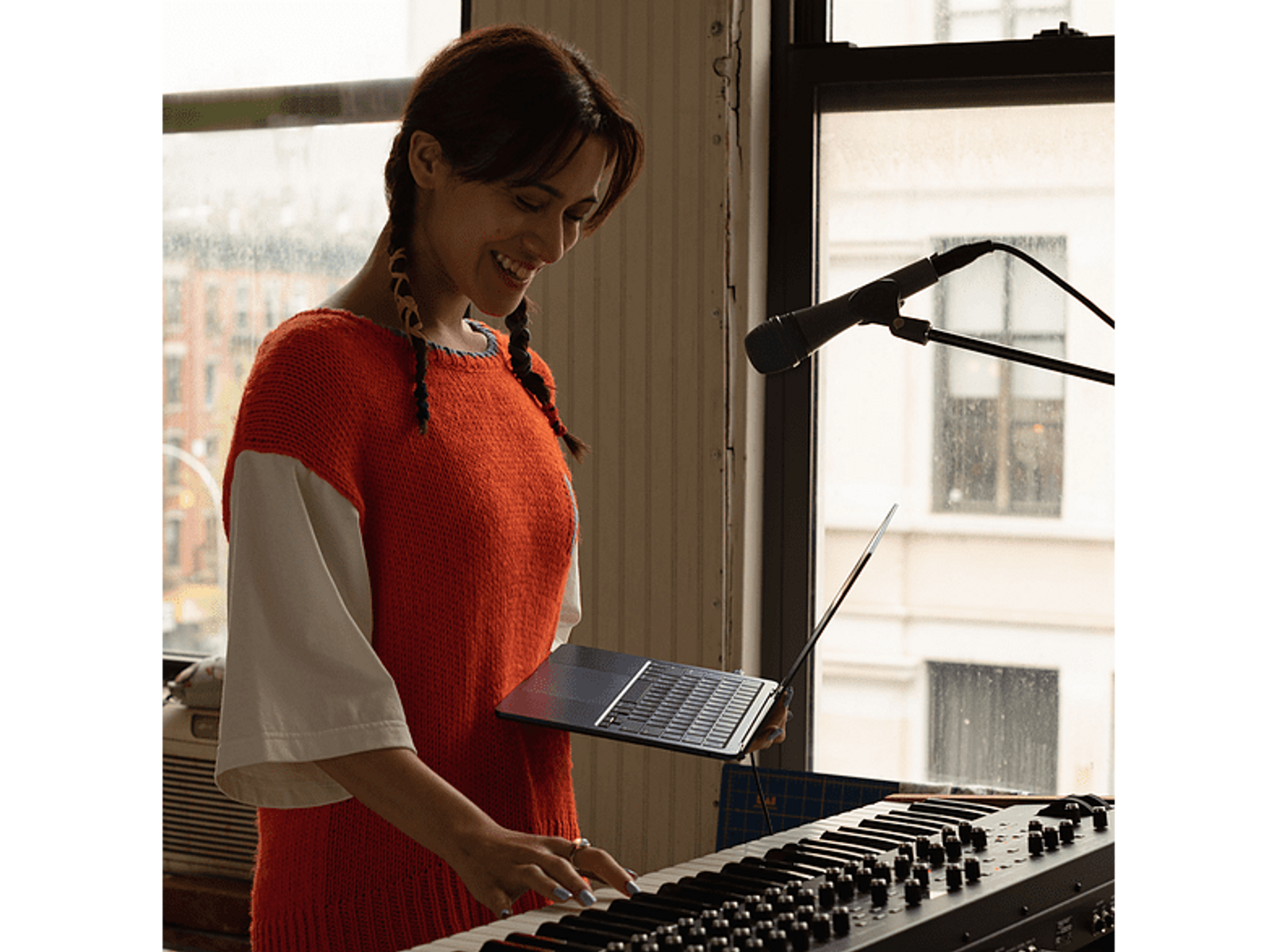 Mujer tocando el teclado, sonriendo, con un suéter naranja, con micrófono y portátil.