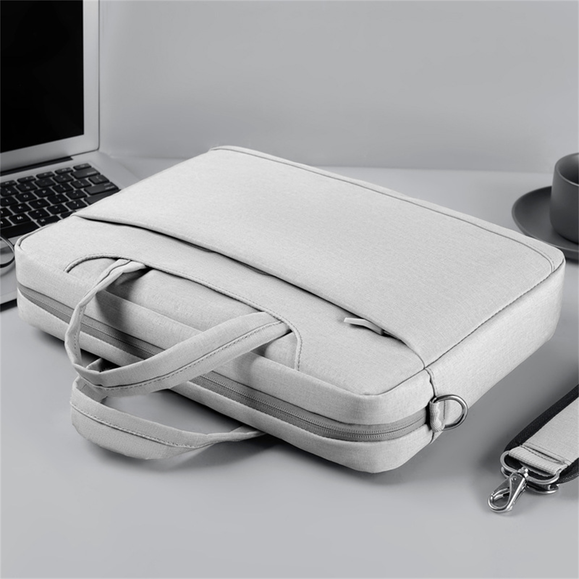 Graue Laptoptasche auf hellgrauer Oberfläche. Laptop dahinter, Tasse rechts.