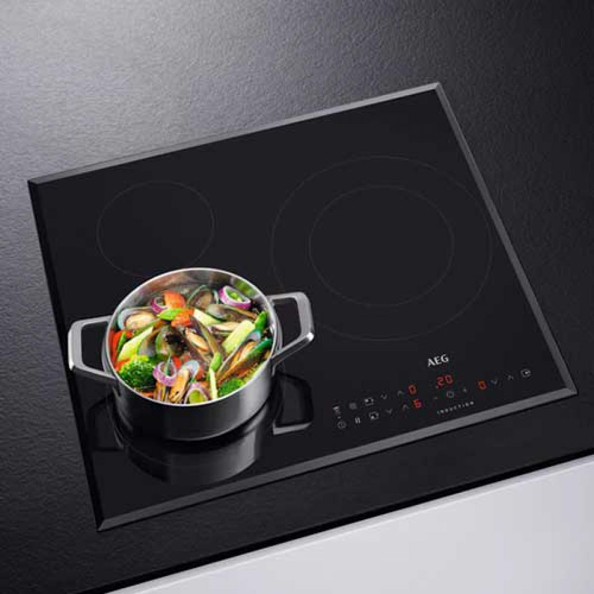 Una encimera negra con dos quemadores. Se coloca una olla con comida. El logo de AEG es visible.