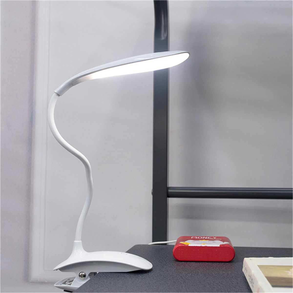 Eine weiße Schreibtischlampe mit gebogenem Hals und Clipfuß. Auf einem Schreibtisch neben roter Box.
