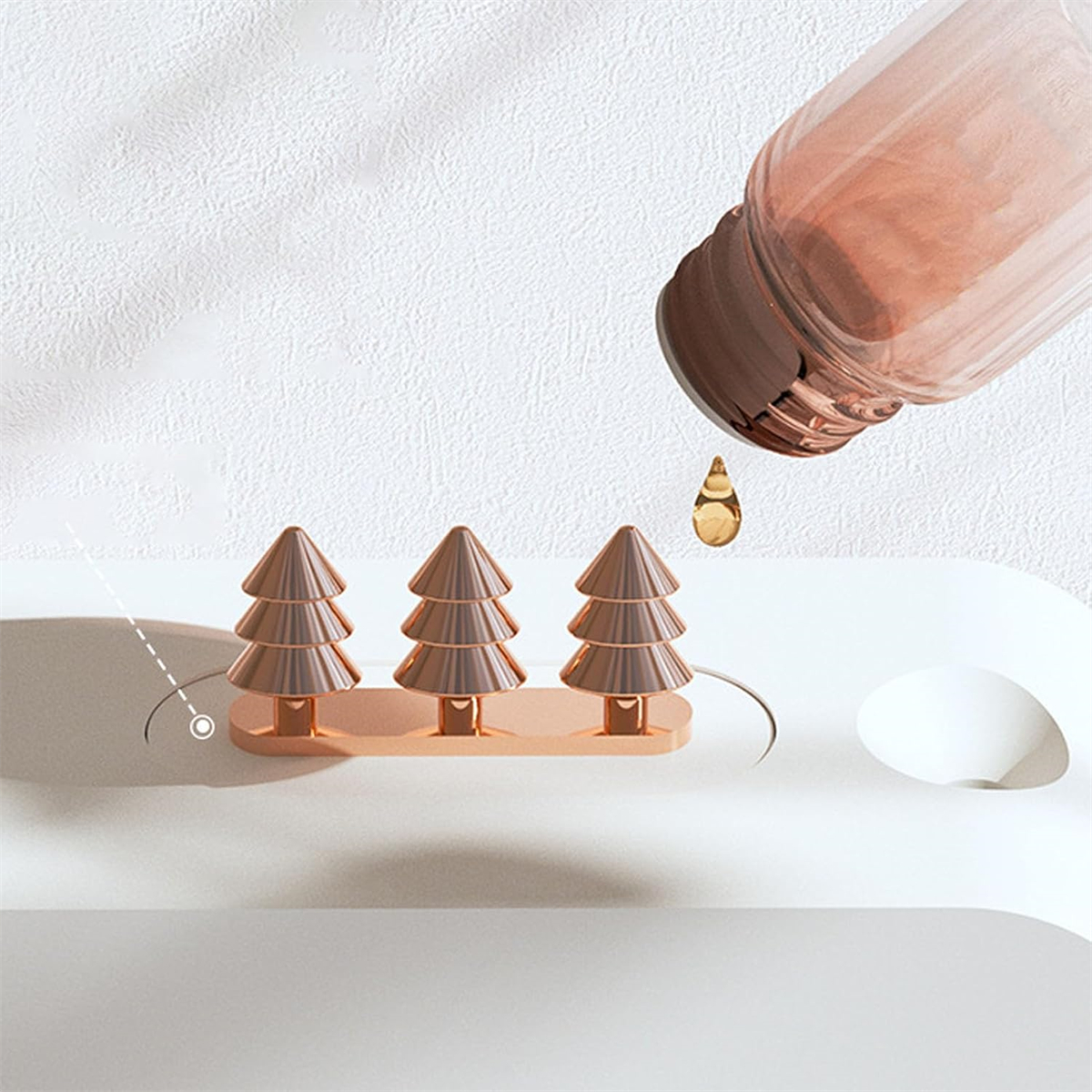 Drei goldene metallische Weihnachtsbäume auf weißer Oberfläche, mit einer Flasche, die Flüssigkeit darauf gießt.