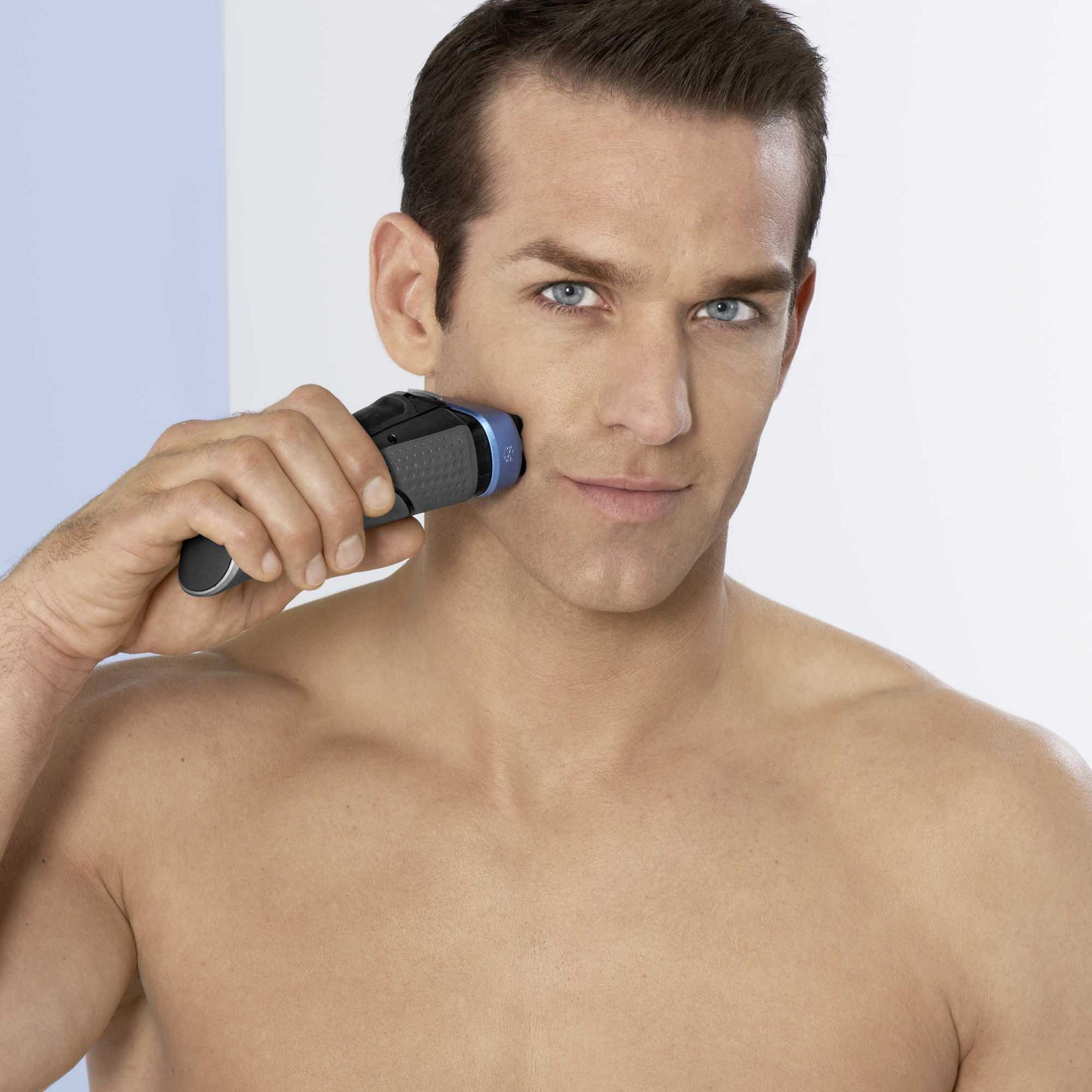 Ein Mann rasiert sich mit einem schwarz-blauen Elektrorasierer. Der Mann hat helle Haut und blaue Augen.