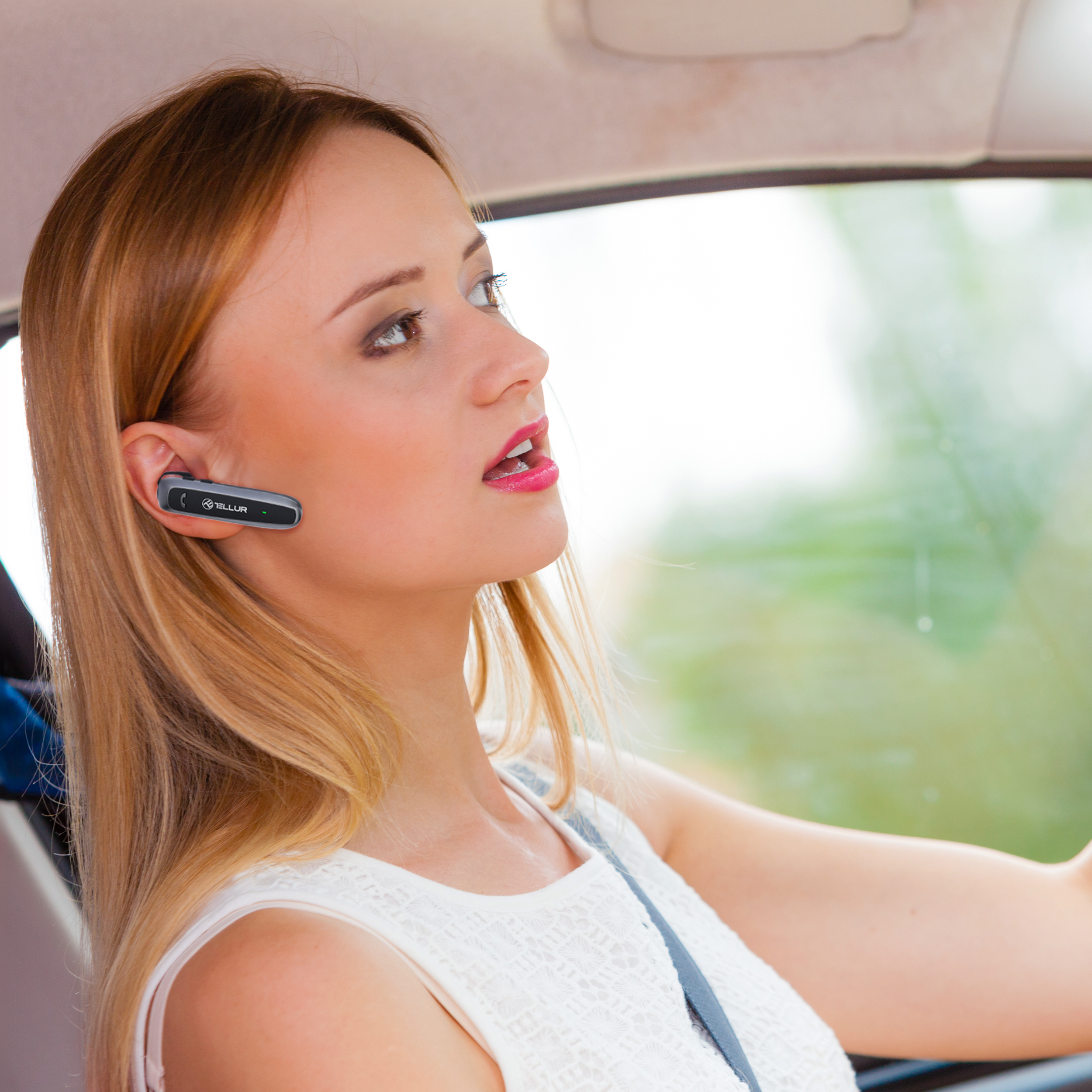 Kobieta w białej sukience prowadzi samochód i ma na sobie niebieską słuchawkę.