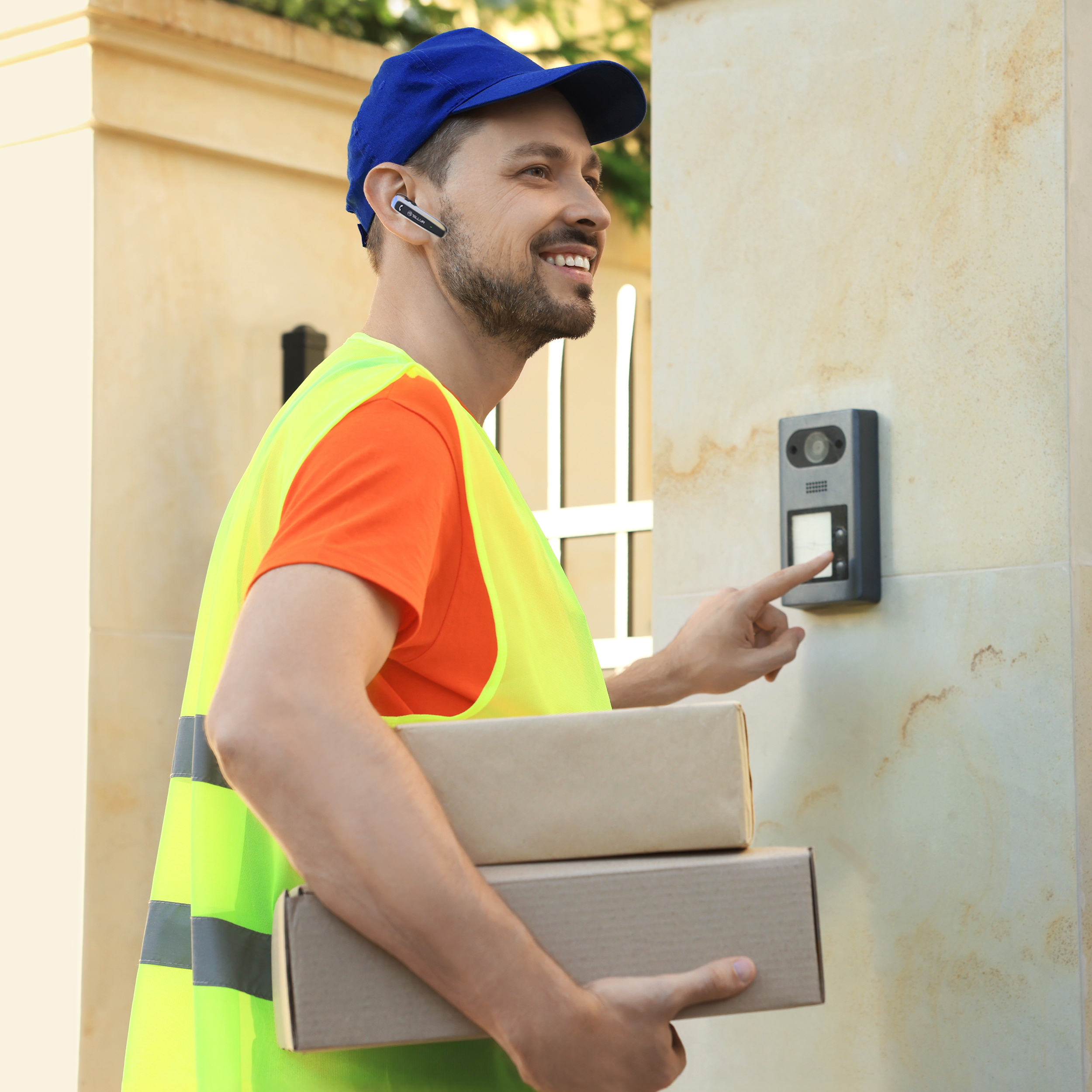Dostawca w niebieskiej czapce i żółtej kamizelce. Trzyma dwa pudełka i dzwoni do drzwi.