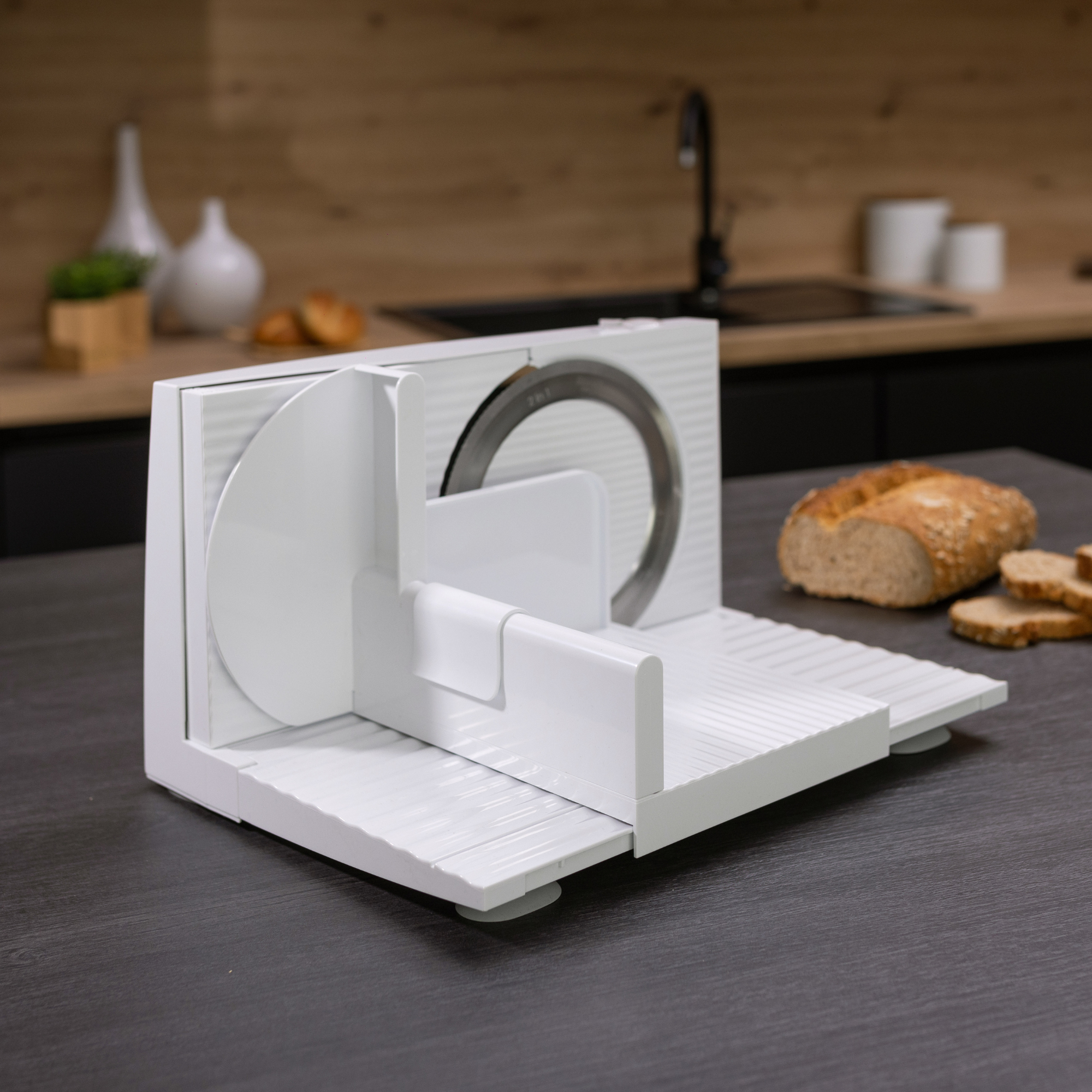 Ein weißer Brotschneider mit silberner Klinge auf dunkler Arbeitsplatte. Im Hintergrund Brot und eine Holzküche.