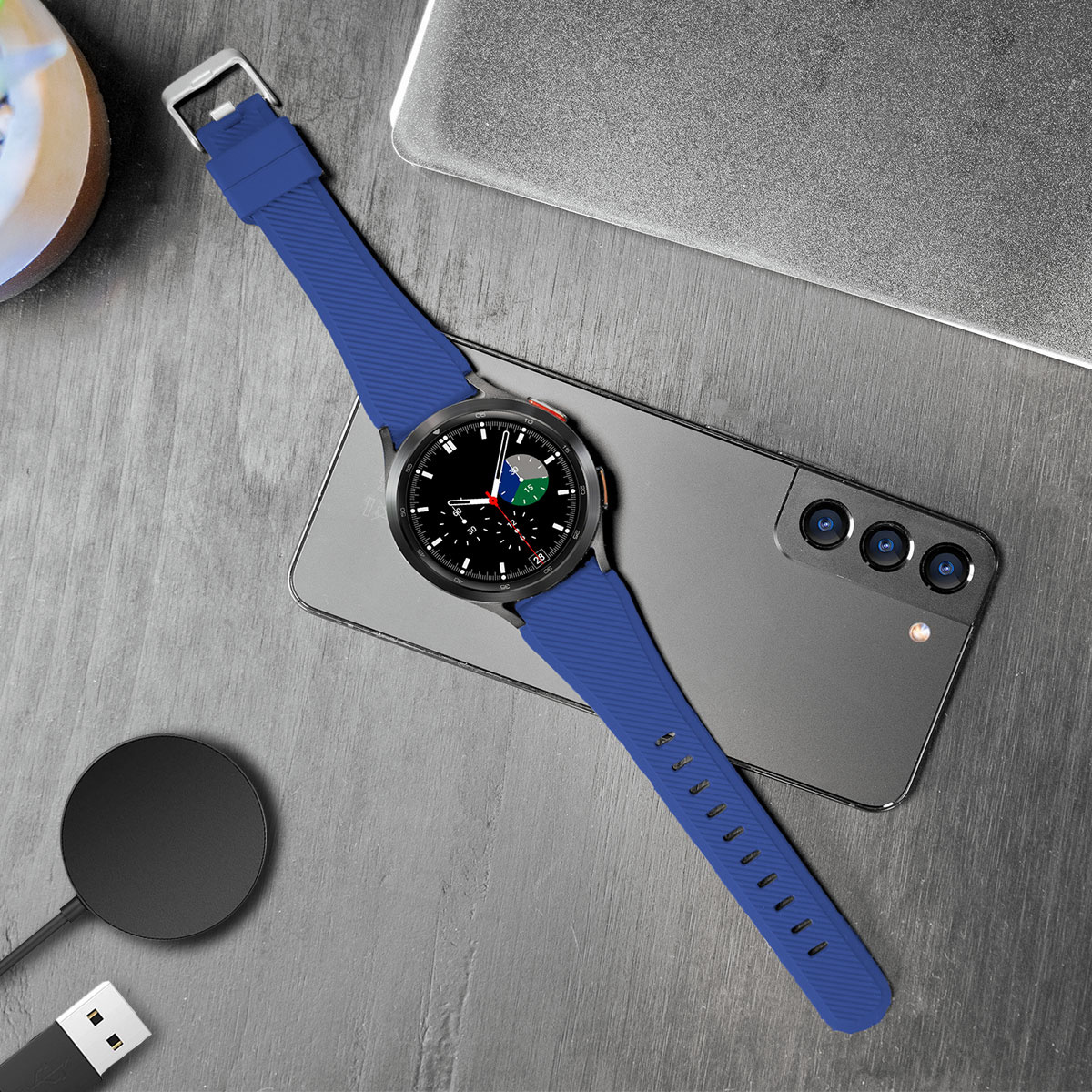Eine schwarze Smartwatch mit blauem Armband, liegt auf einem Telefon neben einer Ladestation und Kabel.