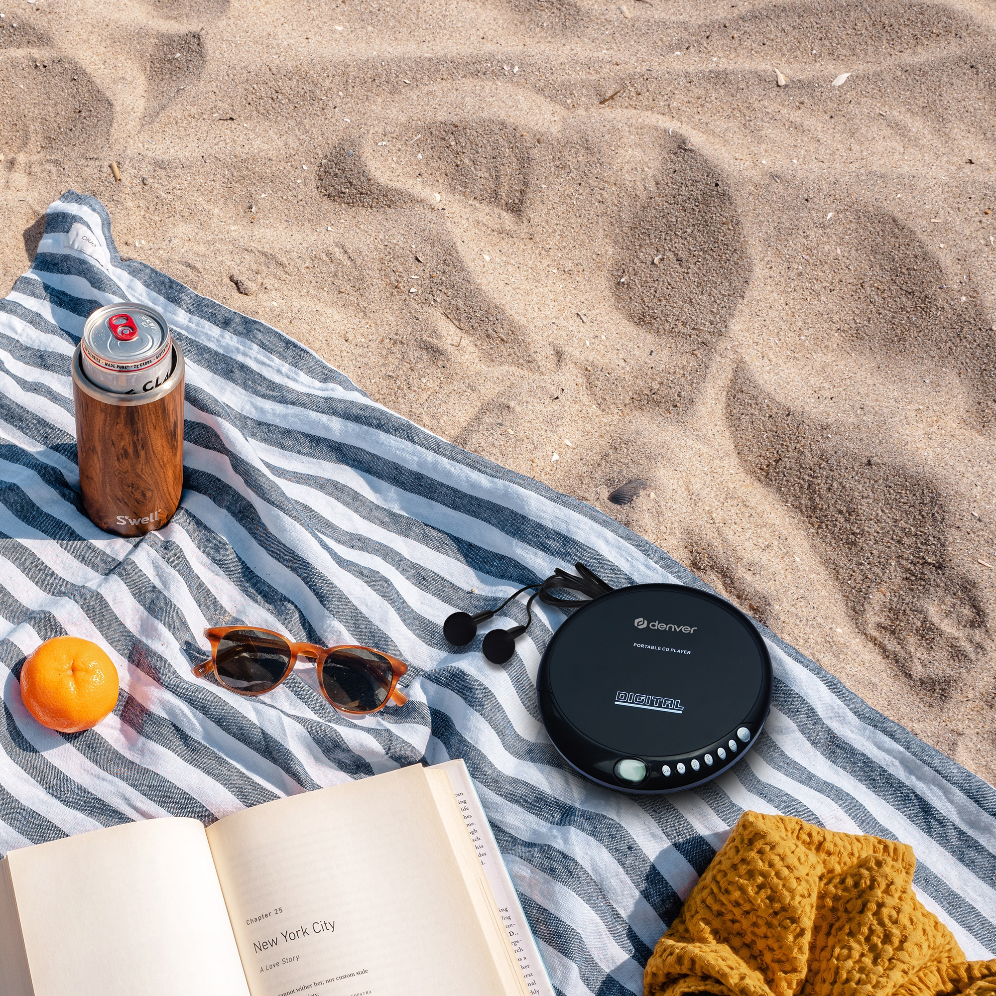 Strandtafereel met een boek, zonnebril, een cd-speler, een drankje en een sinaasappel op een gestreepte handdoek.