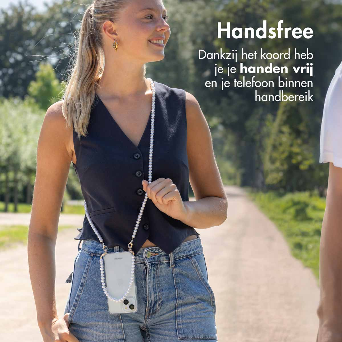 Une femme portant un collier de perles tenant un téléphone. Elle porte un gilet et un jean, souriant.