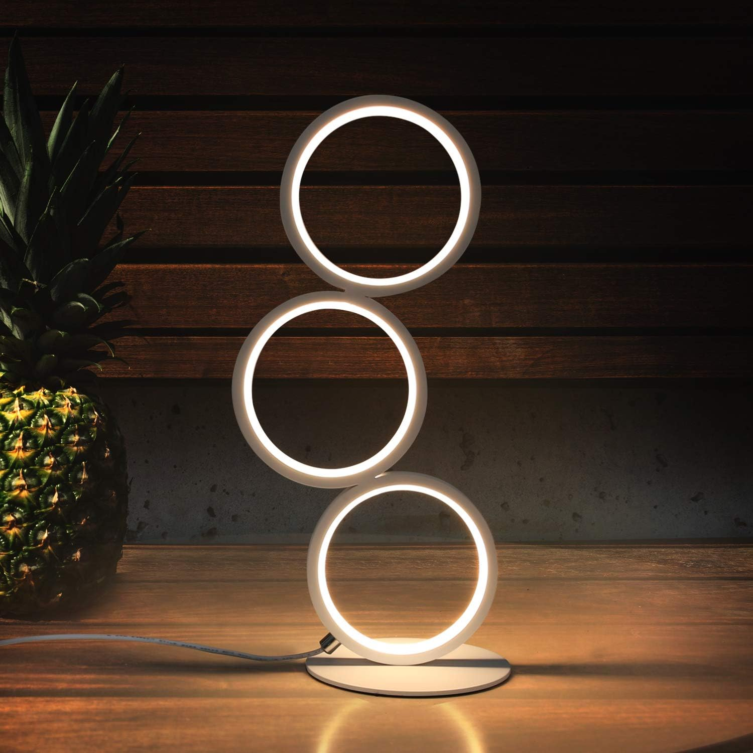 Biała lampa z trzema podświetlanymi pierścieniami. Ananas w tle na drewnianej powierzchni.