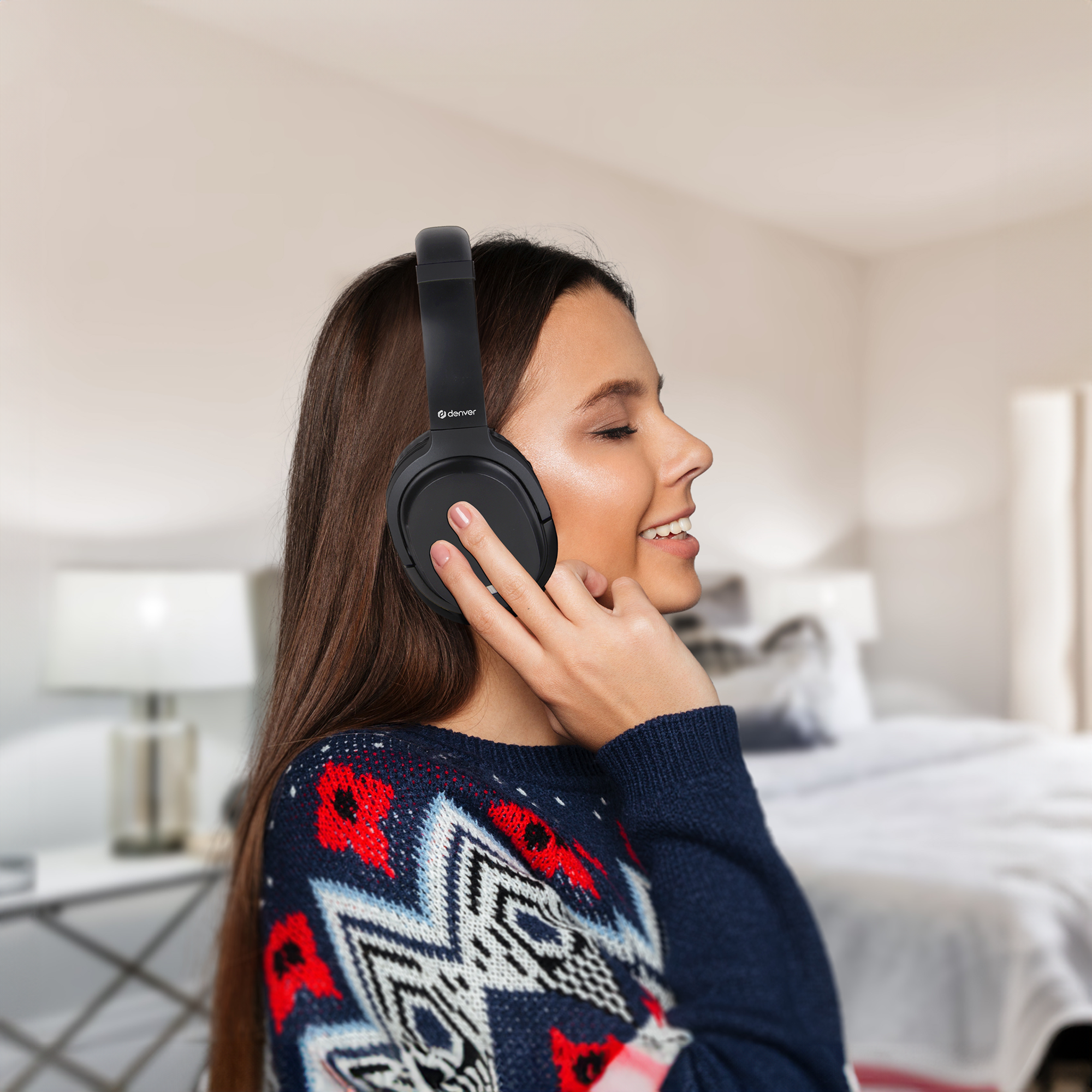 Kobieta w czarnych słuchawkach uśmiecha się. Ma długie brązowe włosy i jest w sypialni.