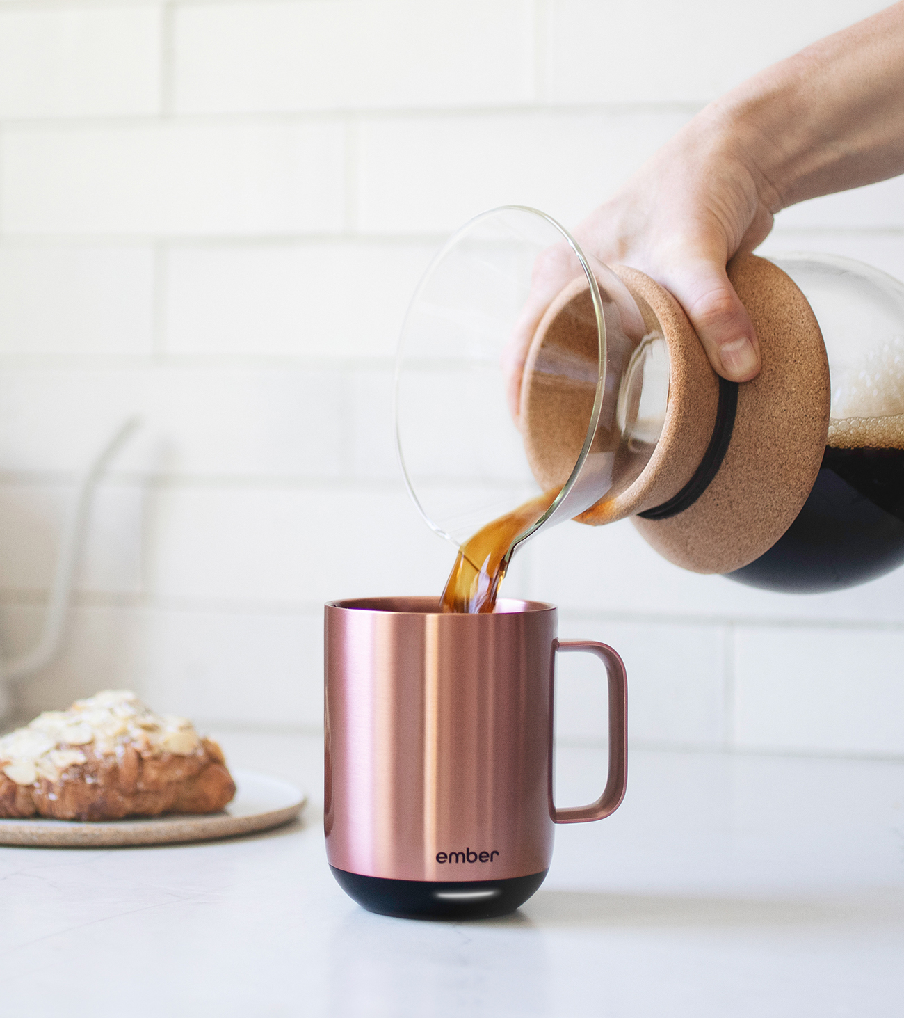 Una persona versa caffè da una caraffa di vetro in una tazza di rame. Un croissant è su un piatto.