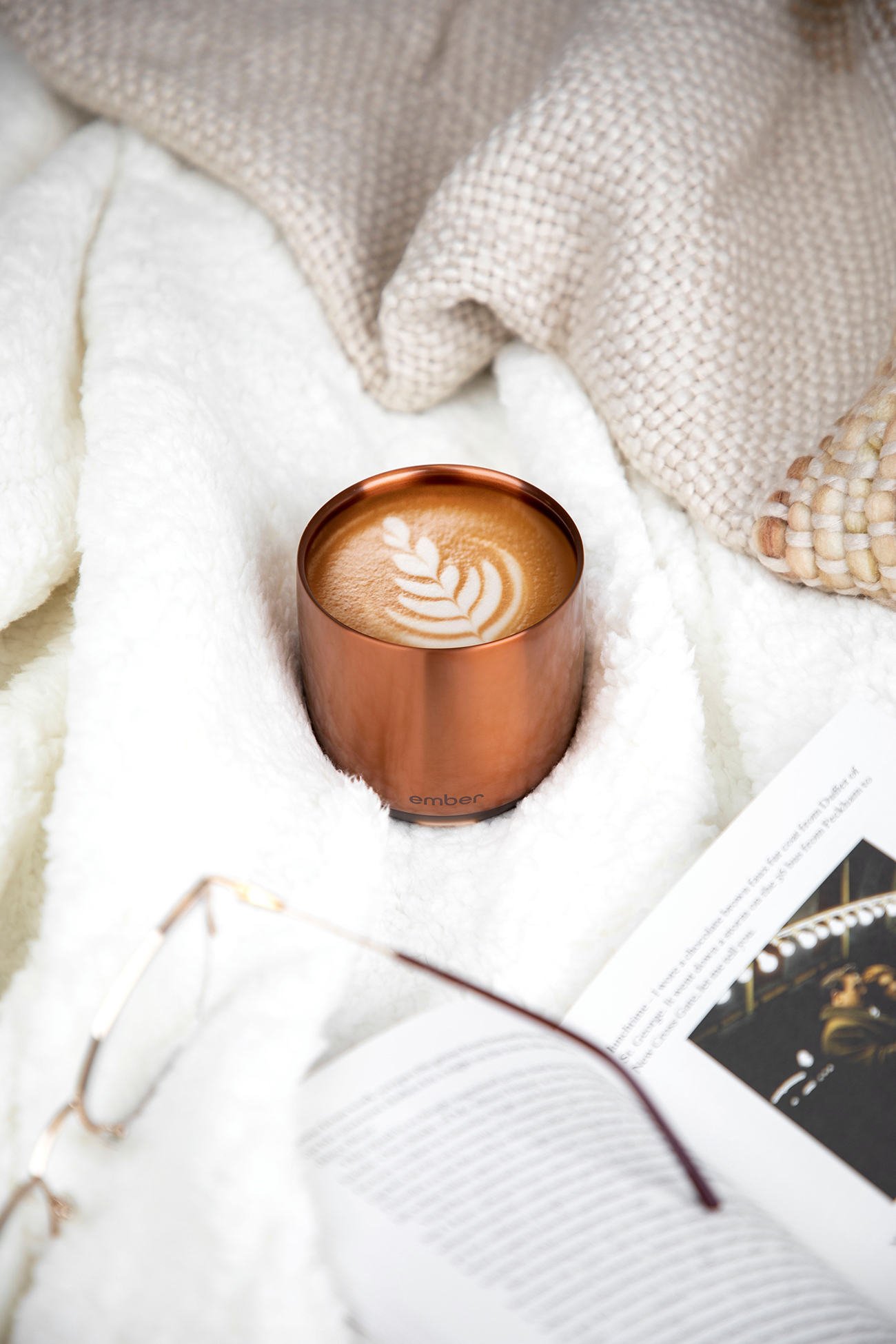 Una tazza di caffè con latte art, su una coperta bianca, con un libro e degli occhiali.