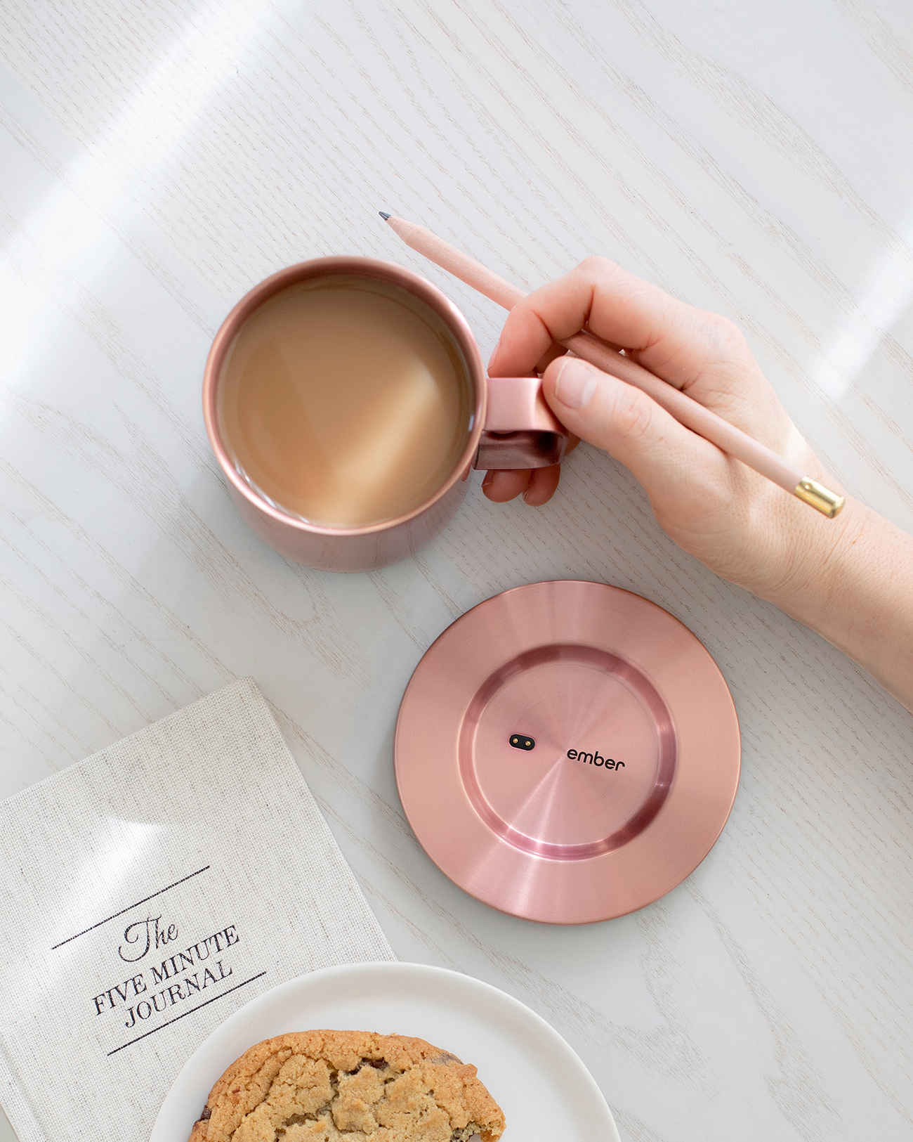 Una tazza rosa con caffè su un sottobicchiere abbinato. Una mano tiene una matita, un diario e un biscotto.