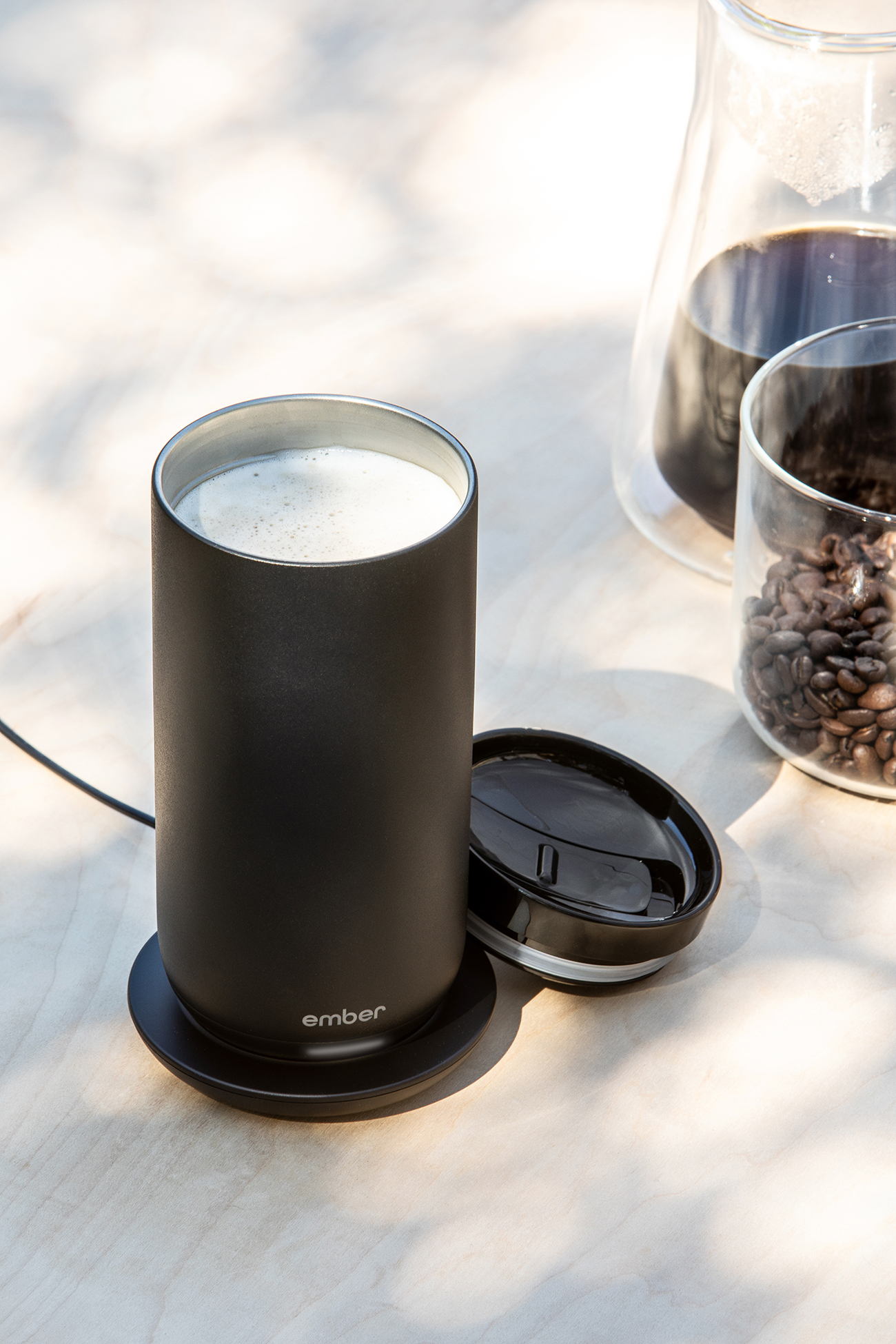 Ein schwarzer Kaffeebecher mit weißem Schaum, auf einem schwarzen Untersetzer. Im Hintergrund eine Glas-Kaffeekanne.