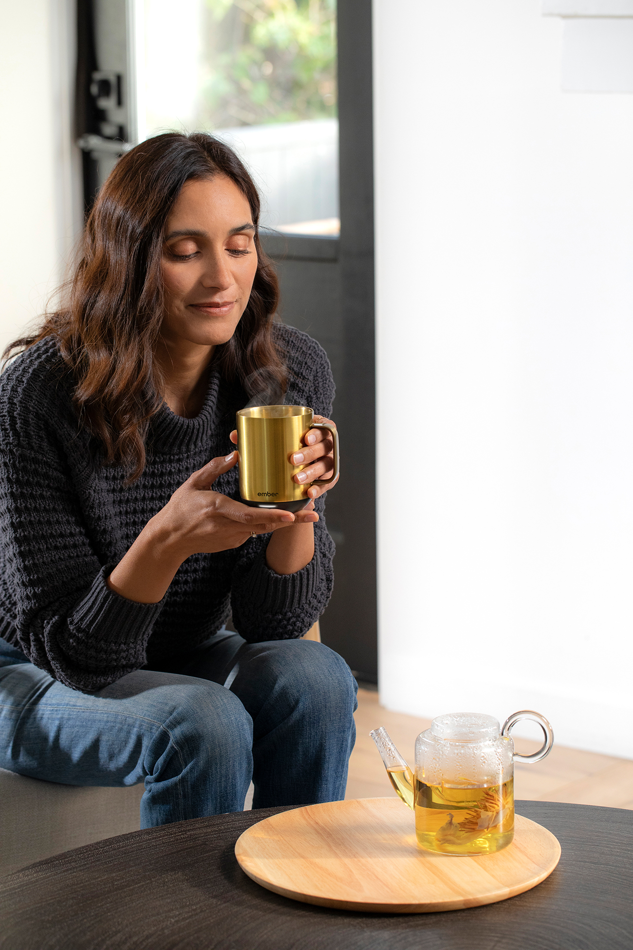 Eine Frau trinkt aus einer goldenen Tasse, mit einer gläsernen Teekanne und einem Holztablett.