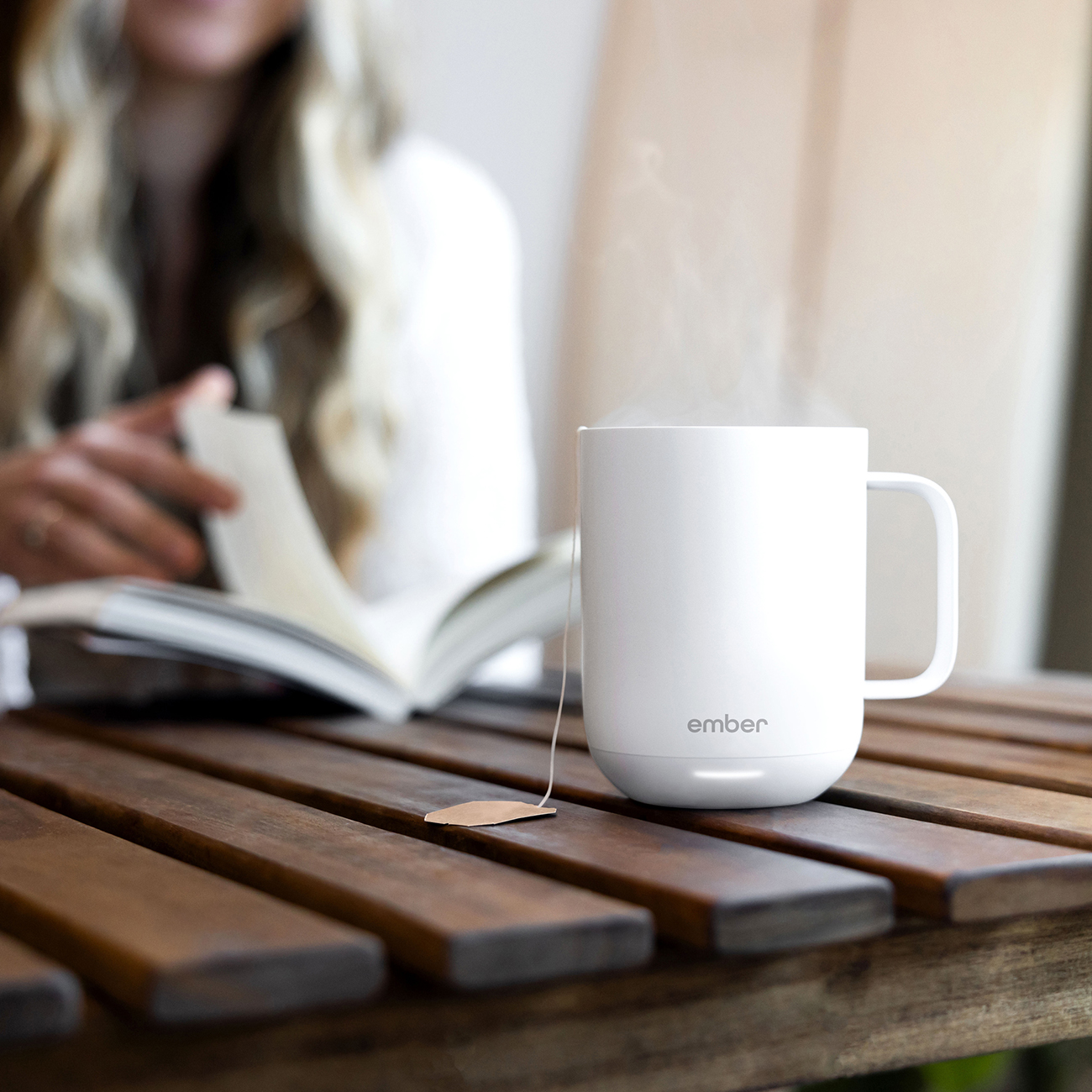 Tazza bianca con vapore, bustina di tè dentro, su tavolo di legno. Sfondo sfocato, donna che legge.